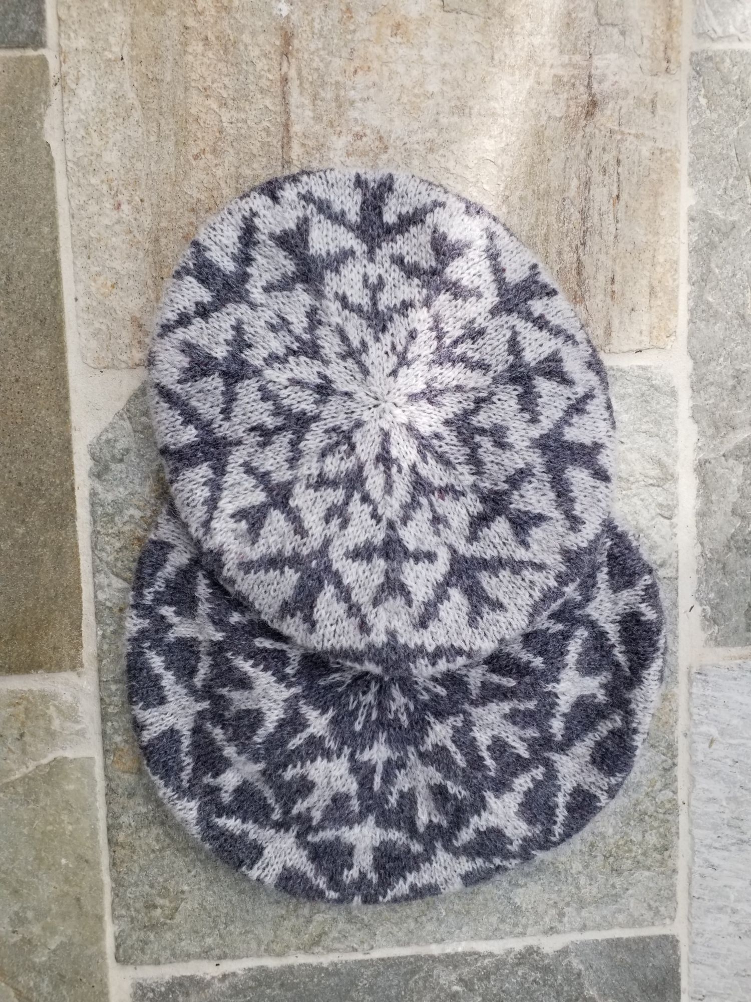 Swifts tam, two samples in grey on white and white on grey. Stranded colourwork motifs of birds flying towards the crown of the hat. The hats are lying on a stone floor.