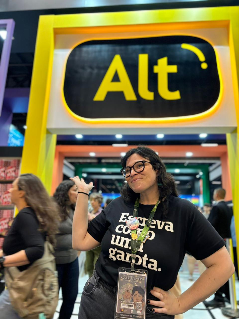 eu, mulher branca, camiseta preta com os dizeres "escreva como uma garota", óculos de grau, cabelo preto, sorrindo na frente do estande da editora Alt na Bienal, na cor amarela.