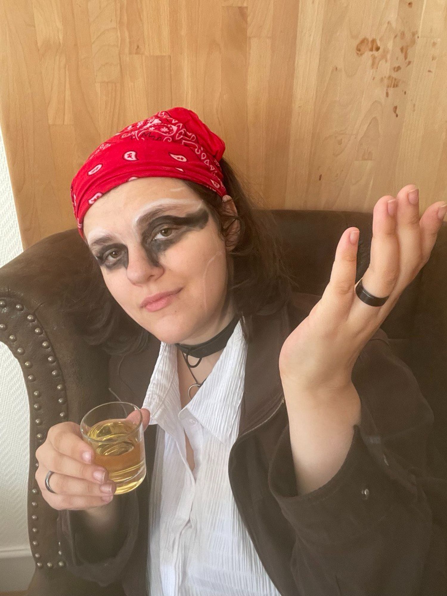 A nonbinary person dressed as a pirate captain with tons of black make up sitting in a leather chair and holding a drink