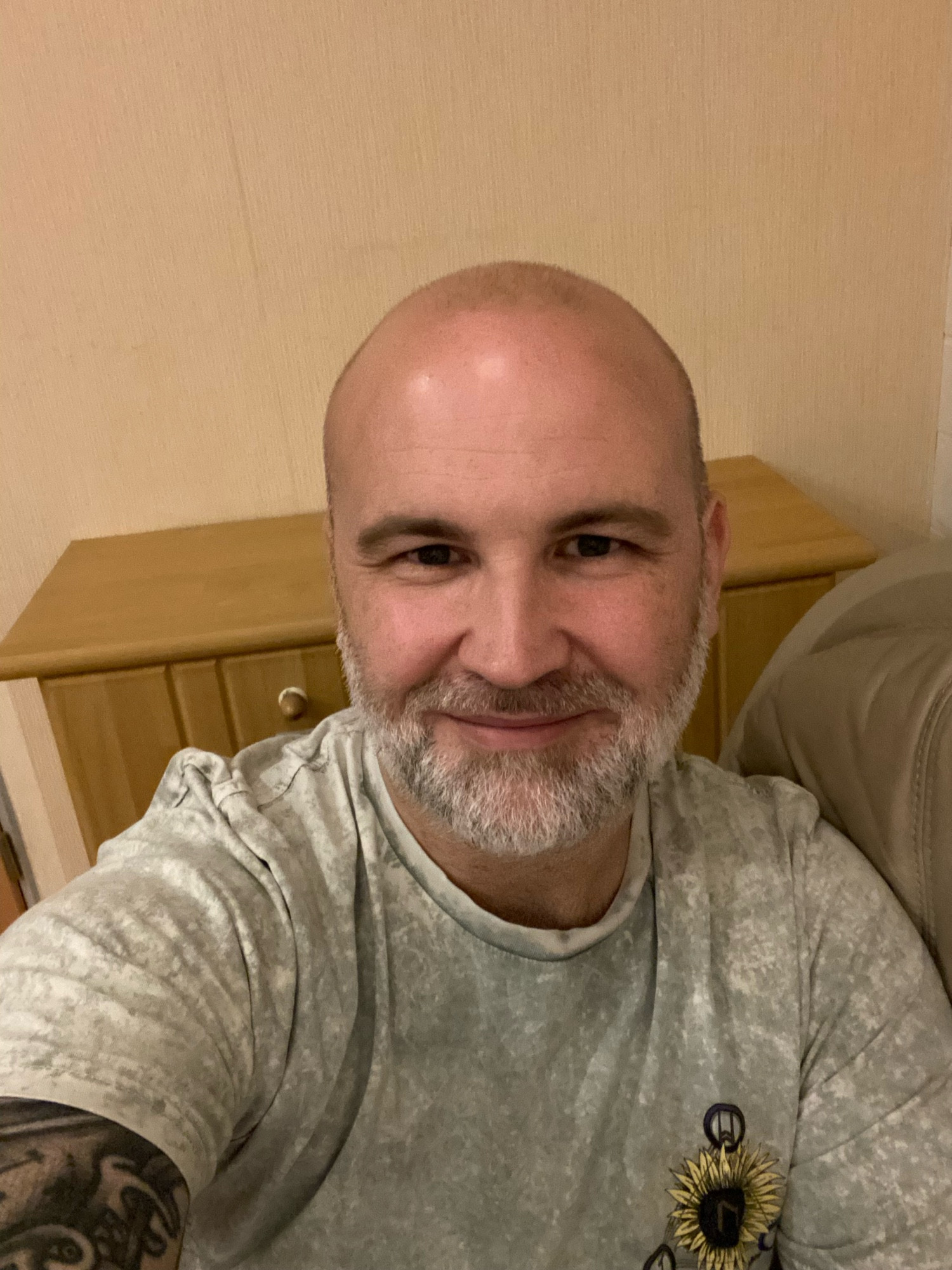 A selfie of me, a middle aged white bald man with a grey beard, smiling and wearing a light grey t shirt with a sunflower print on my left chest. You can see a bit of my right tattoo‘ed Bizeps. I am sitting on a beige leather couch and there is a beech colored sideboard behind me