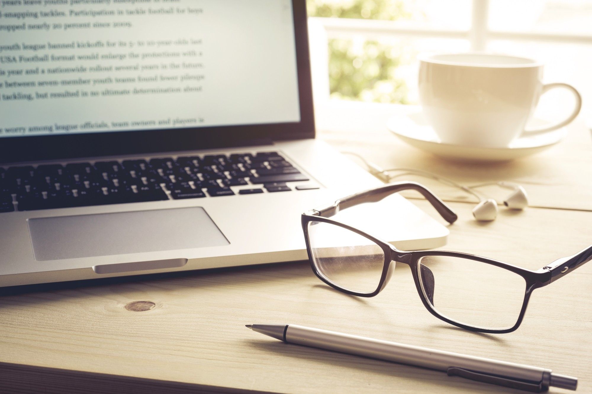 A desk with an open laptop, glasses, a pen, earbuds, and a mug.