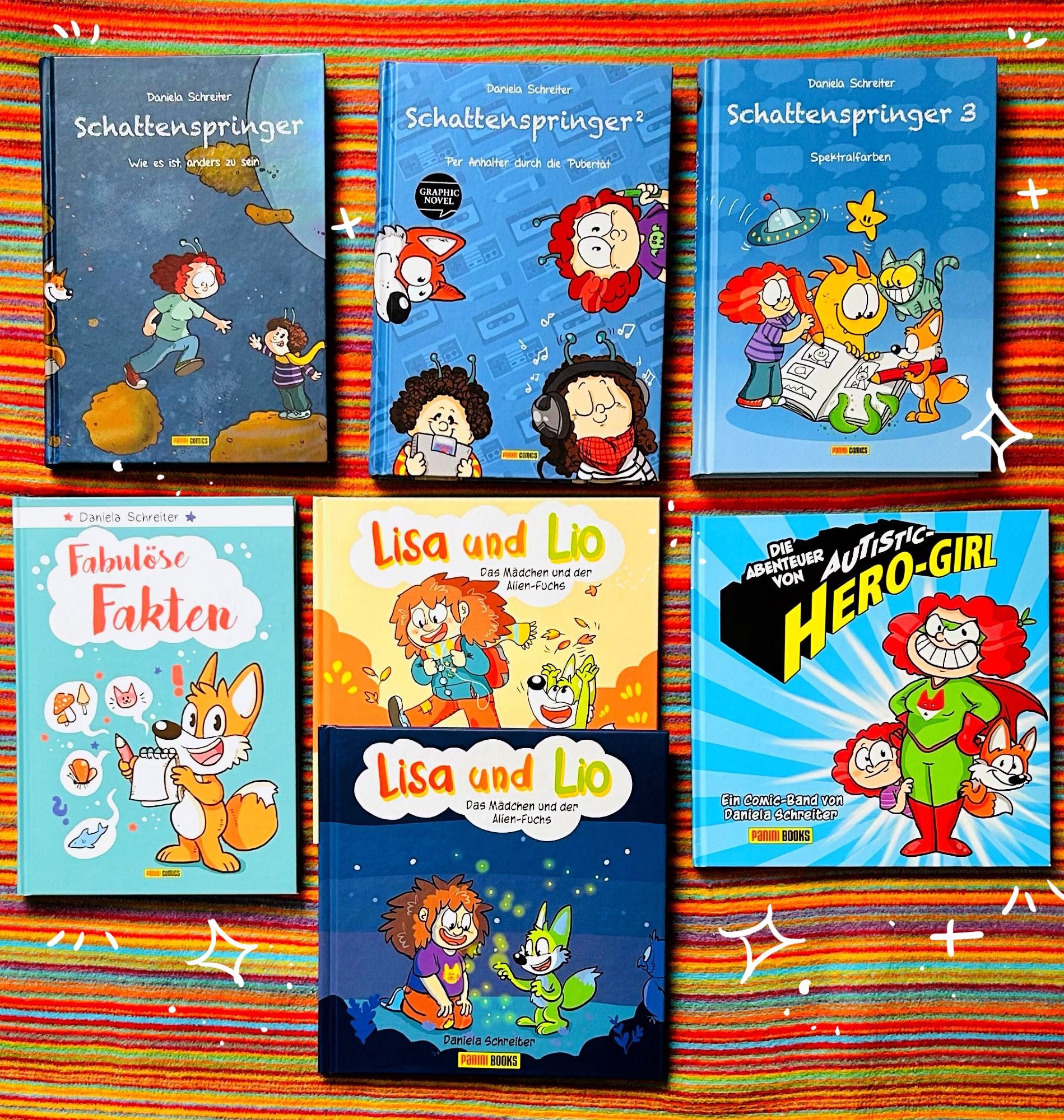 Foto: Sieben Comics sind auf einer buntgestreiften Decke ausgebreitet, die Comics sind Schattenspringer 1 bis 3, Lisa und Lio 1 und 2, Fabulöse Fakten und Autistic Hero-Girl.