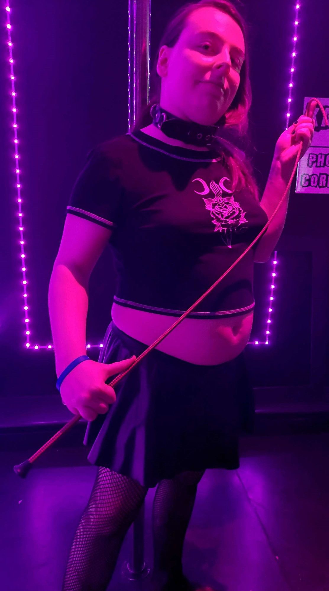 A white trans woman wearing black clothes, a collar, and holding an impact toy called a dressage whip stands in purple lighting