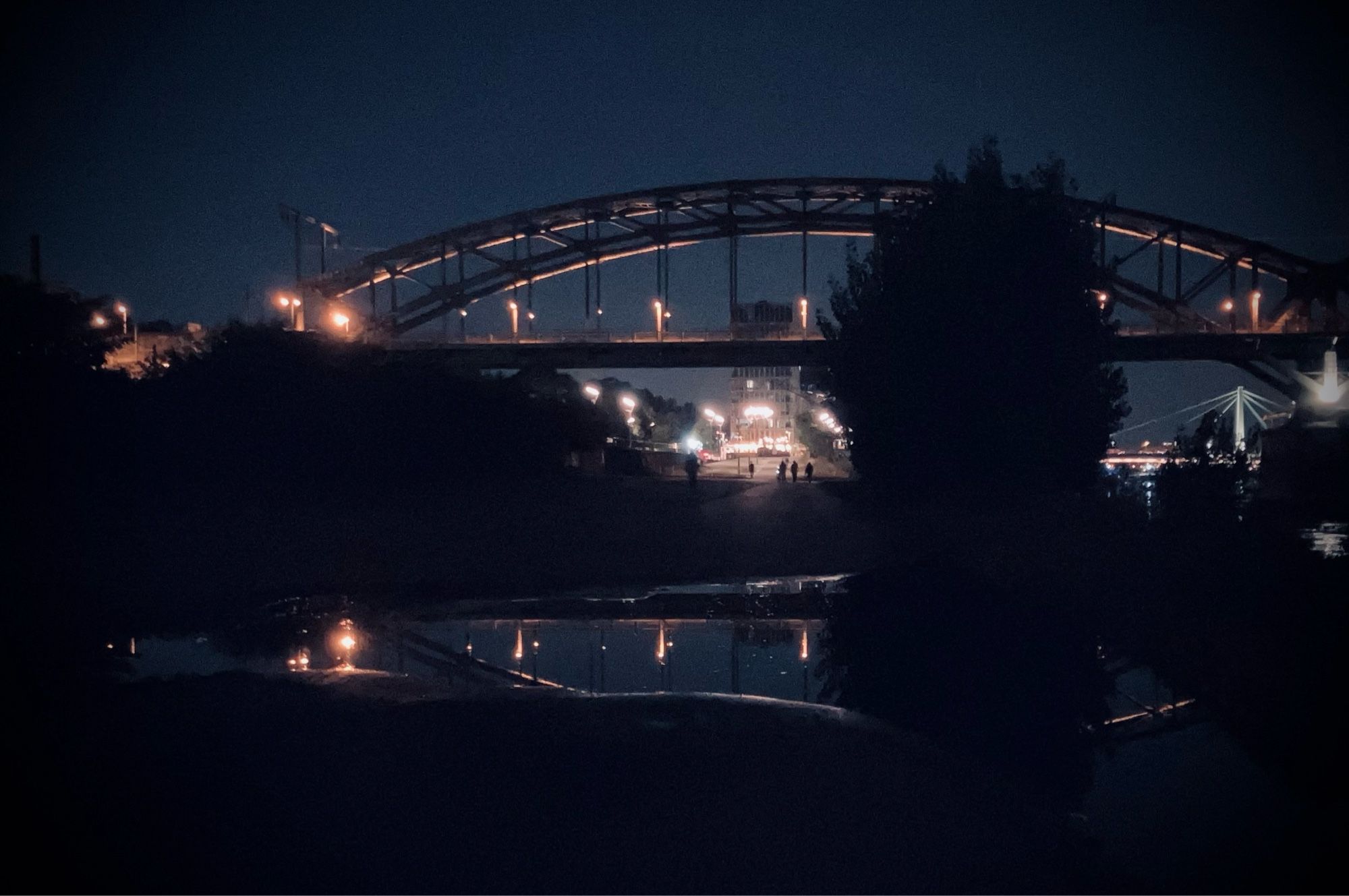 Köln bei Nacht, am Rhein, kurz vor der Südbrücke, die sich teilweise in eine Pfütze spiegelt.