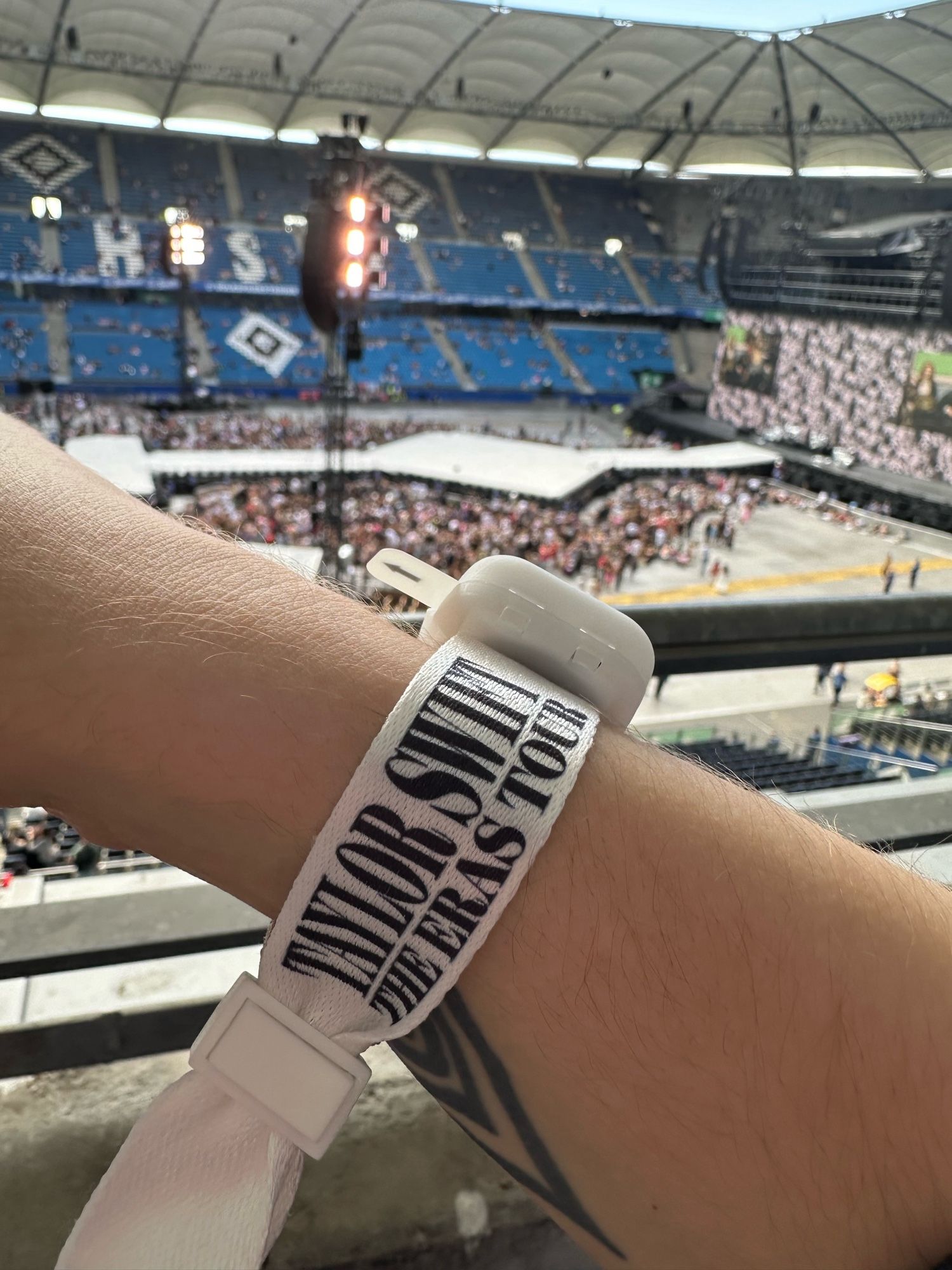 A close-up of a person's wrist wearing a "Taylor Swift The Eras Tour" wristband, with a large stadium and stage setup visible in the background. The stadium is filling with people.