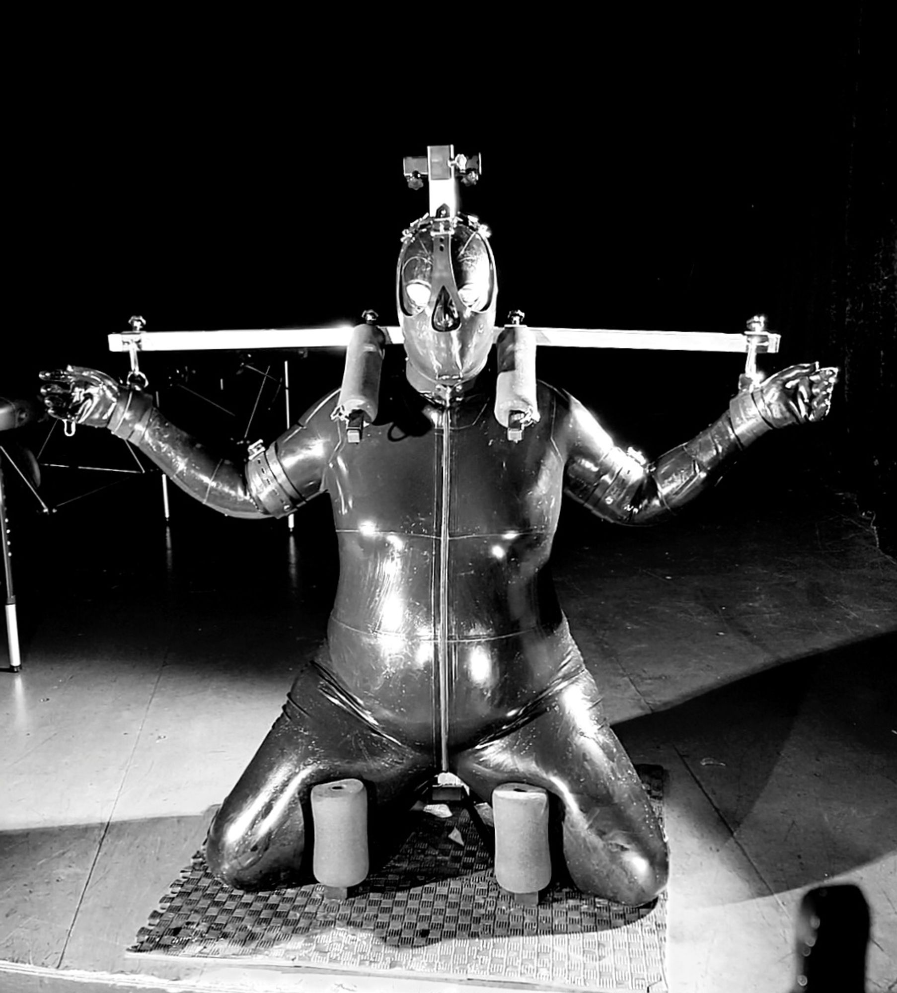 Latex drone locked into the kneeling position in a metal kneeler bondage device with a head harness covering 75% of its skull. Photo in black and white.