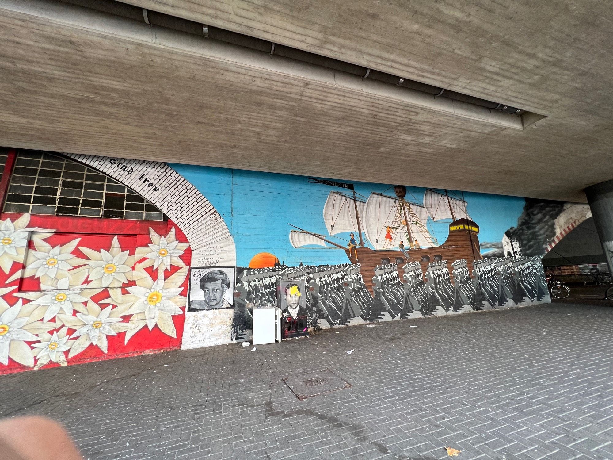 Graffiti aus Köln-Ehrenfeld… “Edelweißpiraten”…

Im linken Drittel des Bildes übergroße Edelweiß-Blüten auf rotem Hintergrund… nach rechts hin das Porträt einer weiblichen Person… im restlichen rechten Bereich Hunderte von im Gleichschritt marschierenden Soldaten (in s/w und grautönen)… im linken Bereich davor ein Porträt eines Mannes… hinter der gesamten Truppe ein blauer Himmel und ein Schiff mit weißen Segeln, auf dem bunt gekleidete junge Menschen sitzen/klettern usw. …