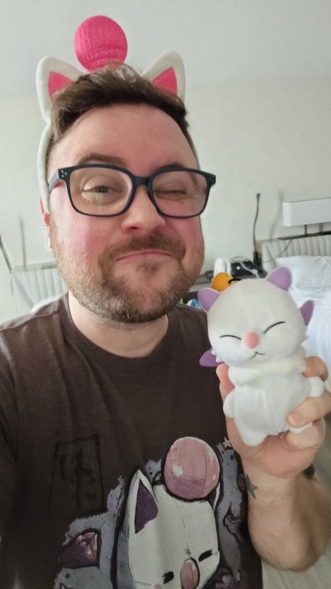 Brandon winking and making a good grin while wearing his moogle ears, a moogle tshirt, and his Mog earrings. He's also holding a tiny moogle plush that turns into a bag