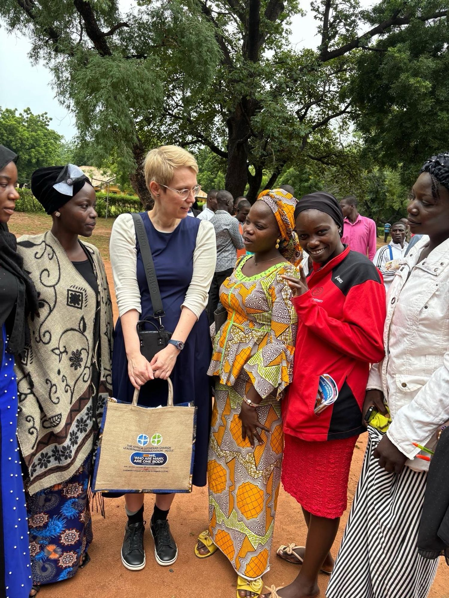 Gruppenphoto - sechs Frauen stehen zusammen im Gespräch, fünf von ihnen sind nigerianische Theologiestudentinnen und ich bin die sechste. Die Studentinnen tragen farbig gemusterte, maßgeschneiderte Kleider und haben passende Tücher um den Kopf gelegt, oder geschlungen. Im Hintergrund sind hohe, alte Bäume voll grünem Laub. Wir stehen auf dem Platz vor der Kapelle des Brønnum Lutheran Seminary. Die Erde unter unseren Füßen ist ocker-orange.