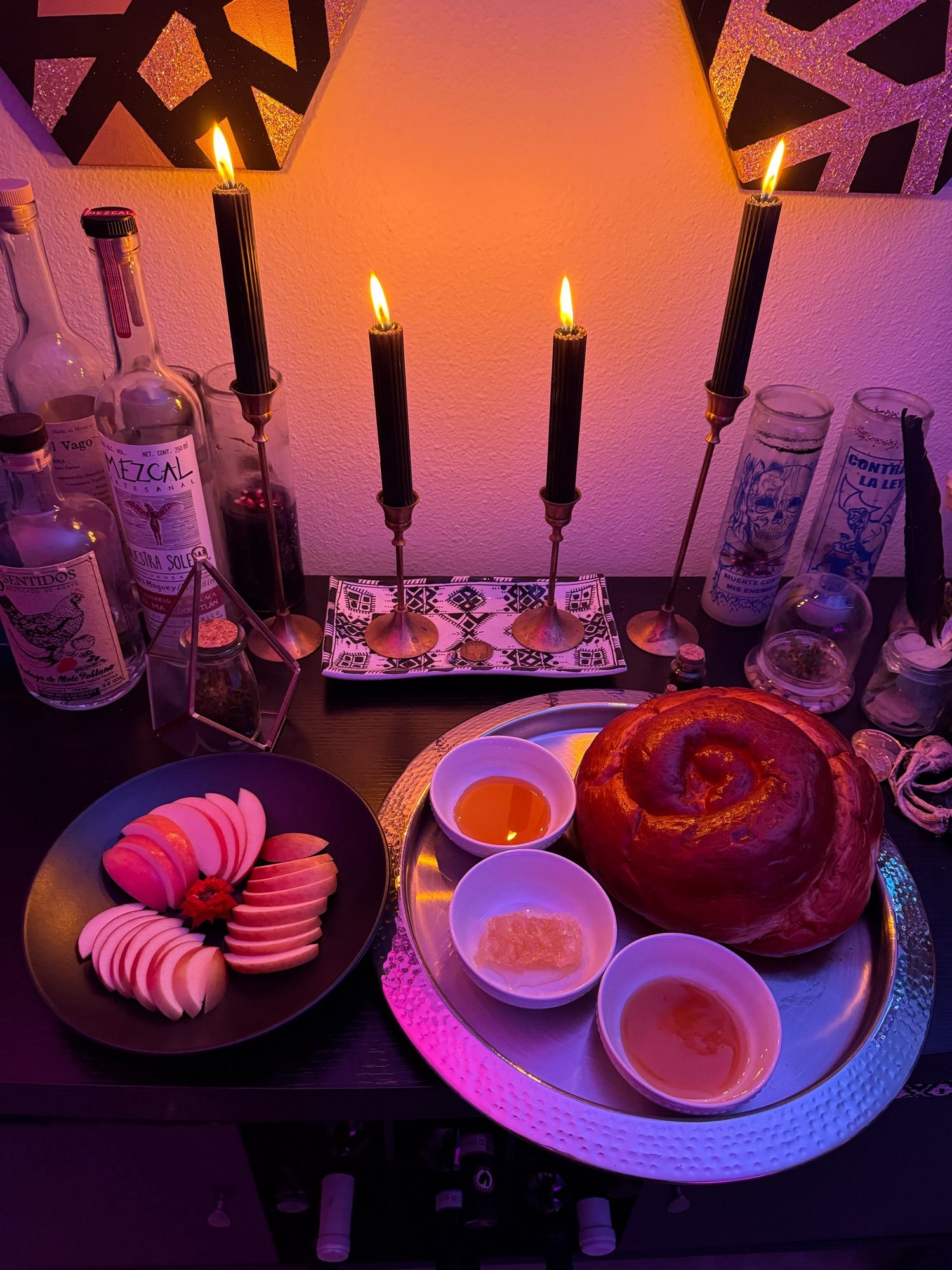 a round loaf of challah sits on a round silver tray with three small dishes of different honeys. beside it is a black plate with three types of apples sliced, and garnished with a tiny rose in the center. four black candles in brass candleholders bathe everything in warm light.