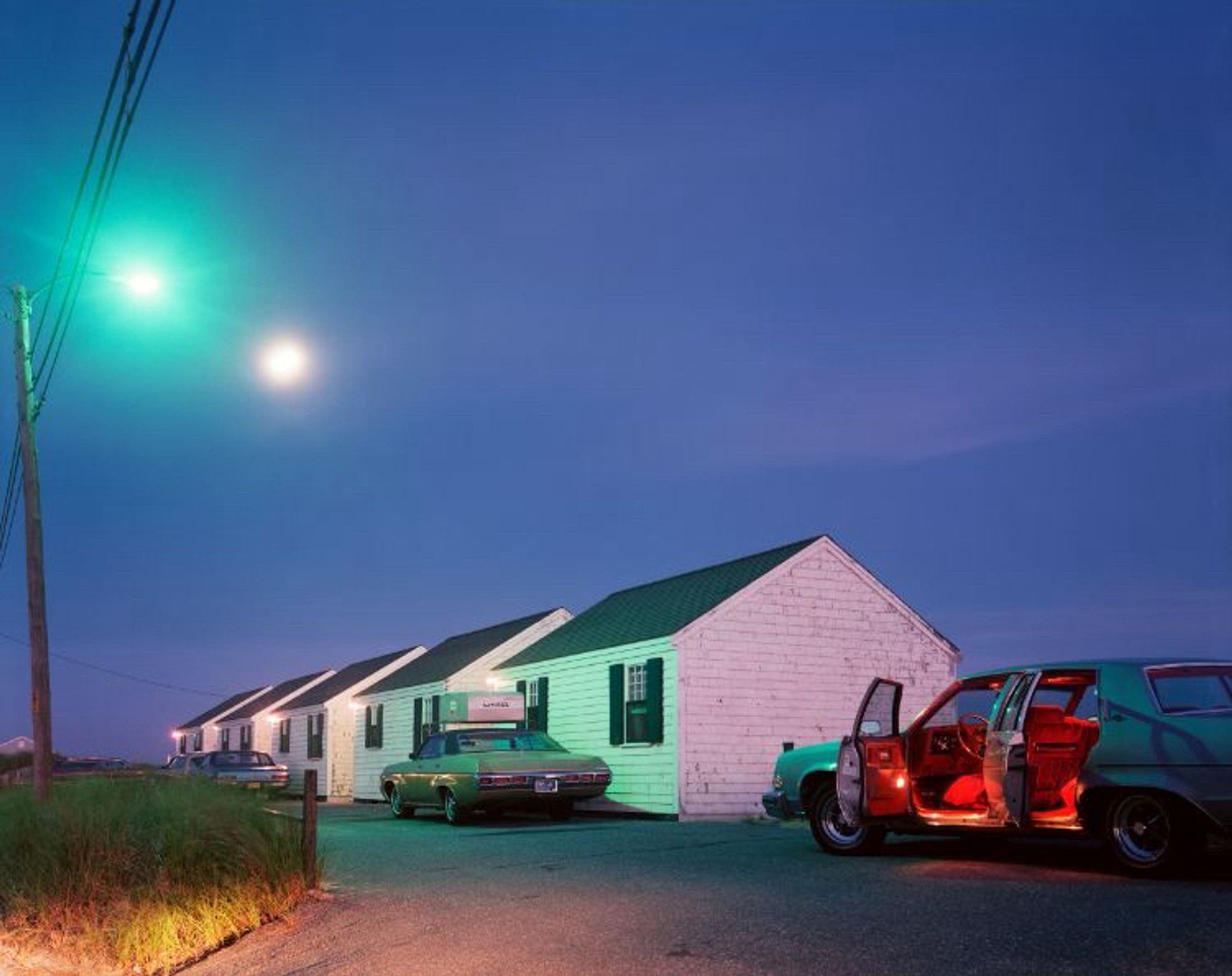 Colour photo of an early evening scene of two cars parked near a white motel building with the nearest car showing red light from the interior through the open doors by Joel Meyerowitz