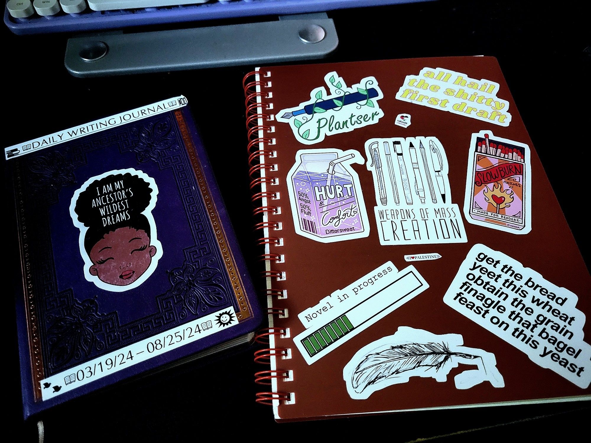 A purple notebook on the left with three stickers: Daily Writing Journal, I Am My Ancestor's Wildest Dreams (over a picture of a black woman smiling).

Red spiral notebook on the right with several motivational stickers: Plantser, All Hail the Shitty First Draft, a picture of various writing instruments labeled Weapons of Mass Creation, a bar half-full of green bars labeled Novel in Progress, a quill dripping ink, and a sticker labeled Get The Bread/Yeet This Wheat/Obtain the Grain/Finagle That Bagel/Feast On This Yeast.