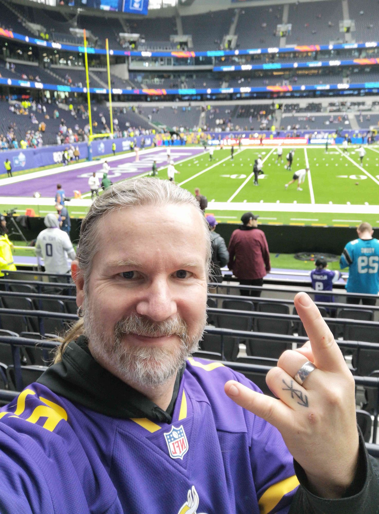 Der User Farbschlurf im Tottenham Stadion. Er trägt ein lila Trikot der Minnesota Vikings und macht mit der rechten Hand die "Pommesgabel". Im Hintergrund sieht man eine der Endzones und einige Spieler beim Aufwärmen.