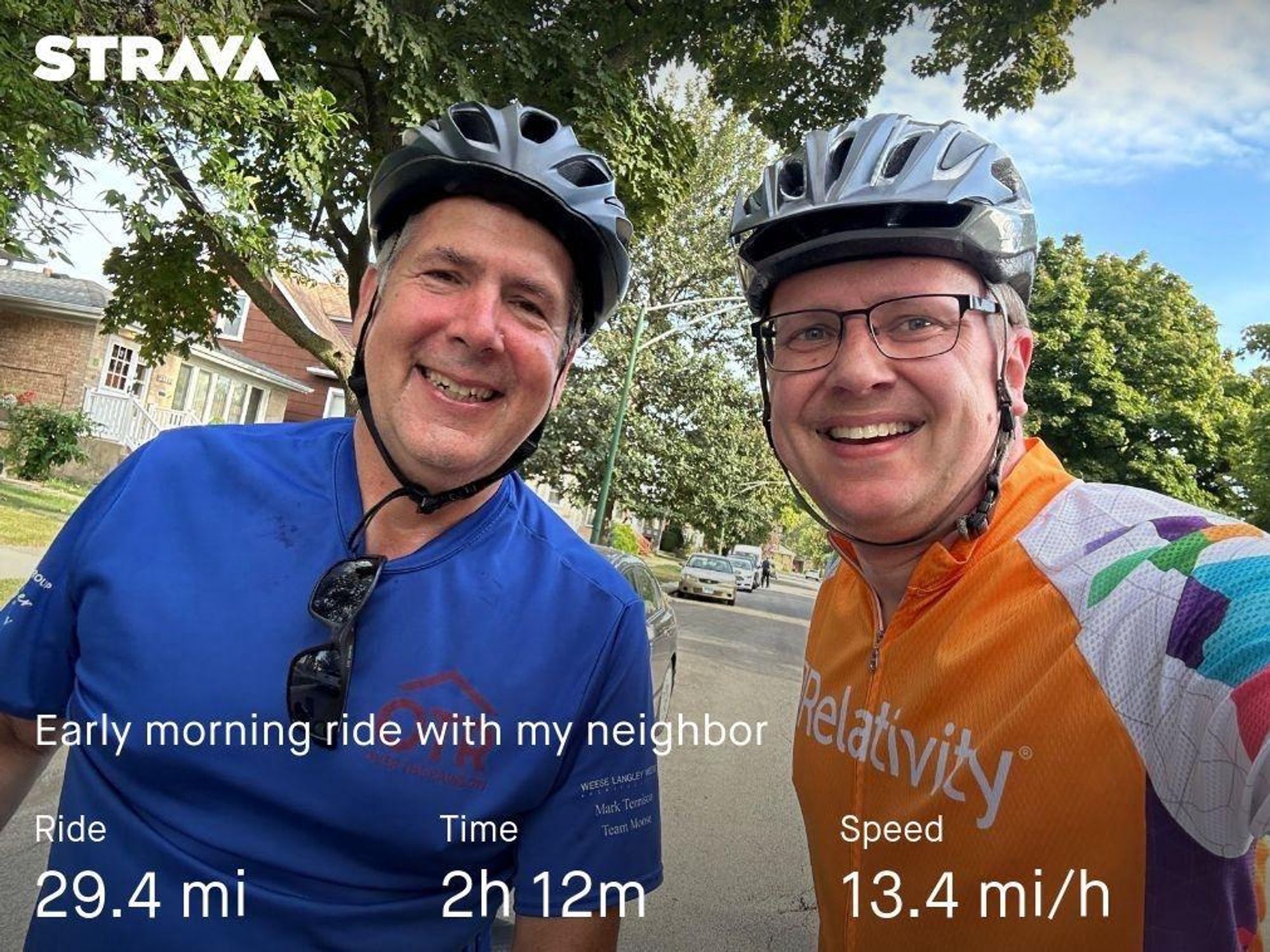 Two cyclists smiling for a selfie during an early morning ride. One is wearing a blue shirt and the other a multicolored jersey. The background shows a neighborhood street with trees and houses. Overlay text indicates the ride distance, time, and speed.