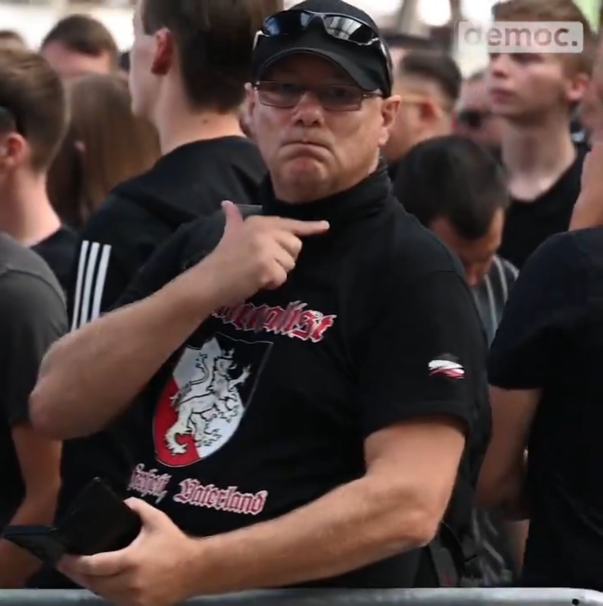 Mann in schwarzem T-Shirt mit Nazi-Symbolik und mit schwarzer Baseballmütze macht eindeutige Geste in Richtung Kameramann (Hals durchschneiden).