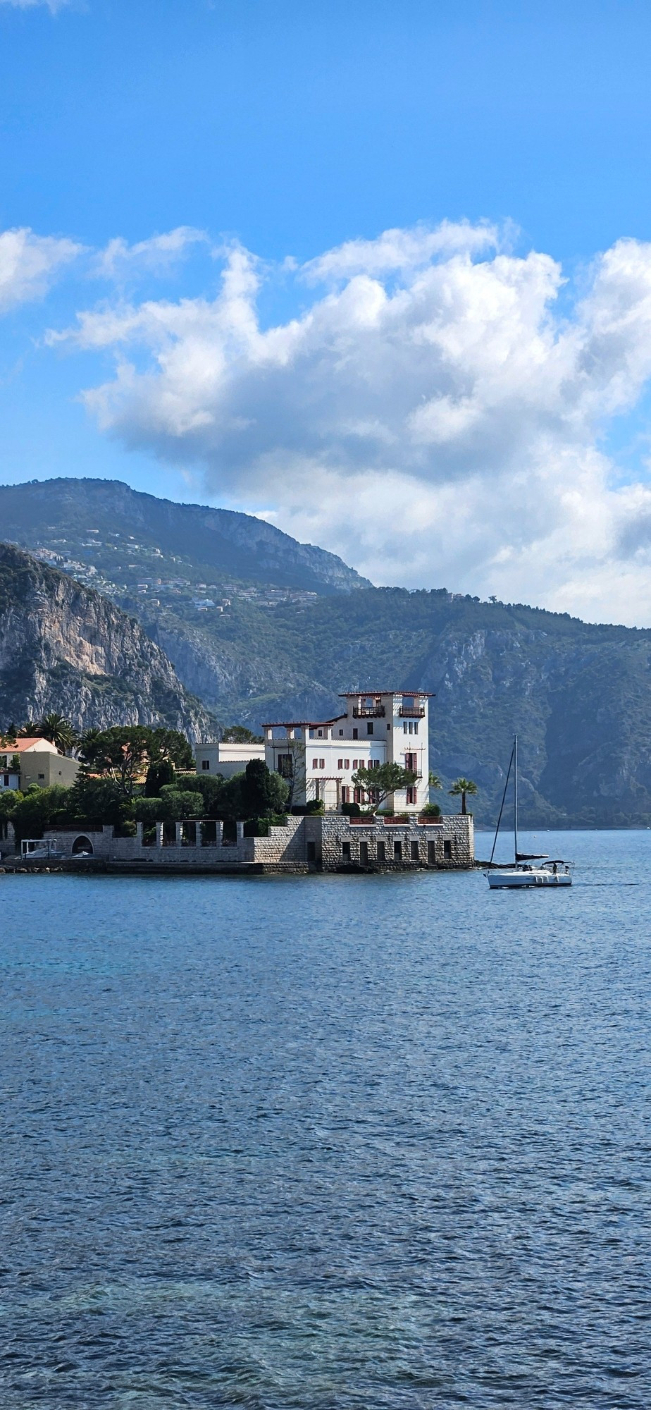 A small blue sailboat glides past a gorgeous villa that stands by the sea. The villa was built in the very early 1900s in an ancient greek revival style. It's been owned by the french state for almost 100 years and is now a museum.