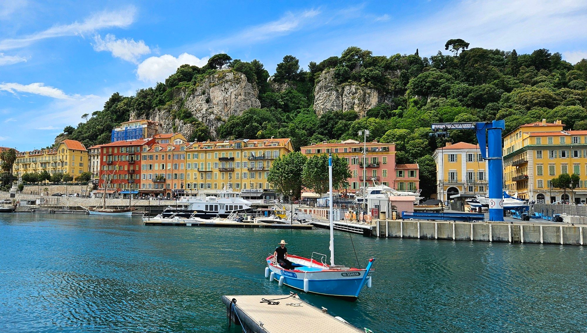 A small boat comes towards the dock, with one person on board. The sailor is wearing black clothes and a strawhat. The boat is painted bright blue, with a red and a white stripe. A row of yellow, pink and red buildings are in the background and a steep cliff, almost covered with trees, looms behind the buildings.