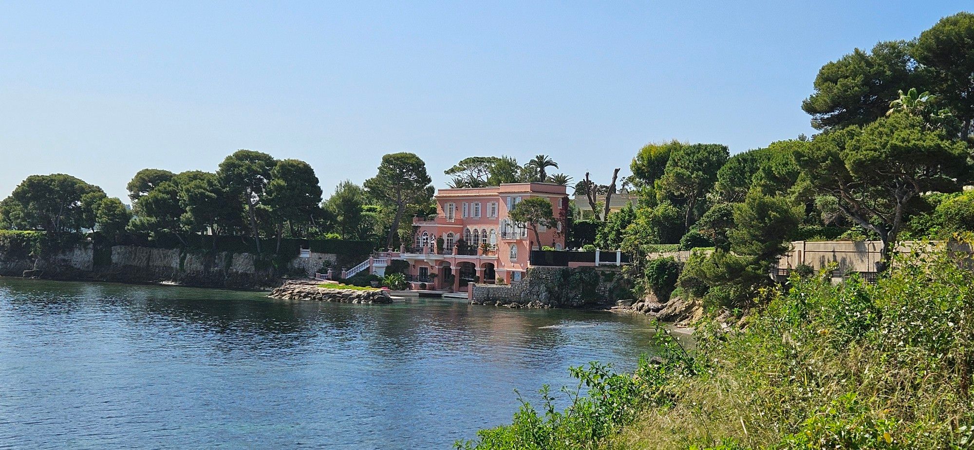 A pink villa by the sea. A small jetty in front of it. Trees line up on both sides of the villa.