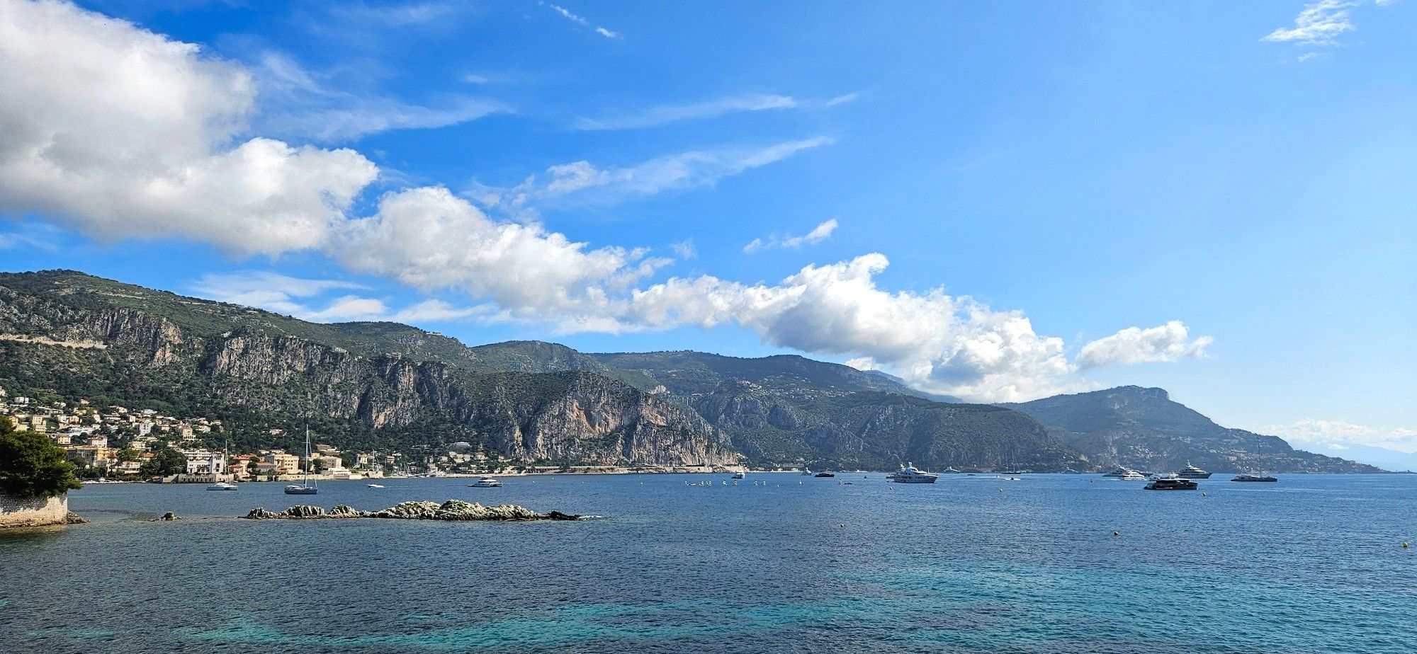 A mountainous coastline, a line of clouds above. A few large yachts and smaller sailboats are scattered around the sea.