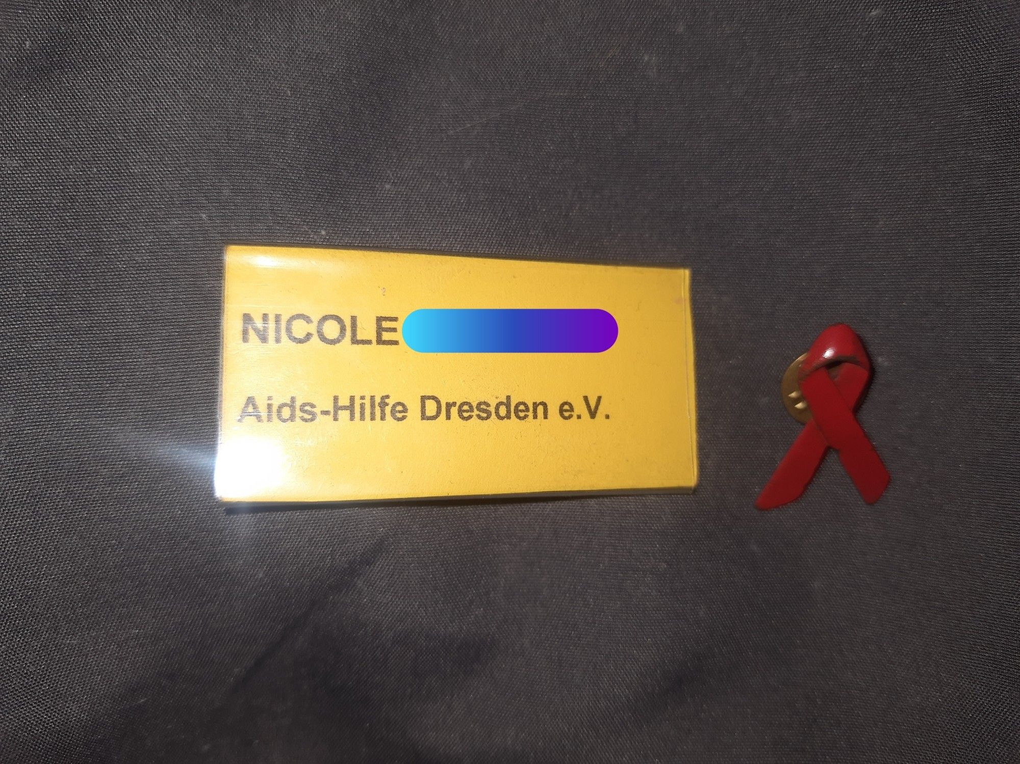 Dunkelgrauer Hintergrund.  Darauf mein Namensschild der Aids- Hilfe Dresden e.V. und die rote Schleife als Pin.