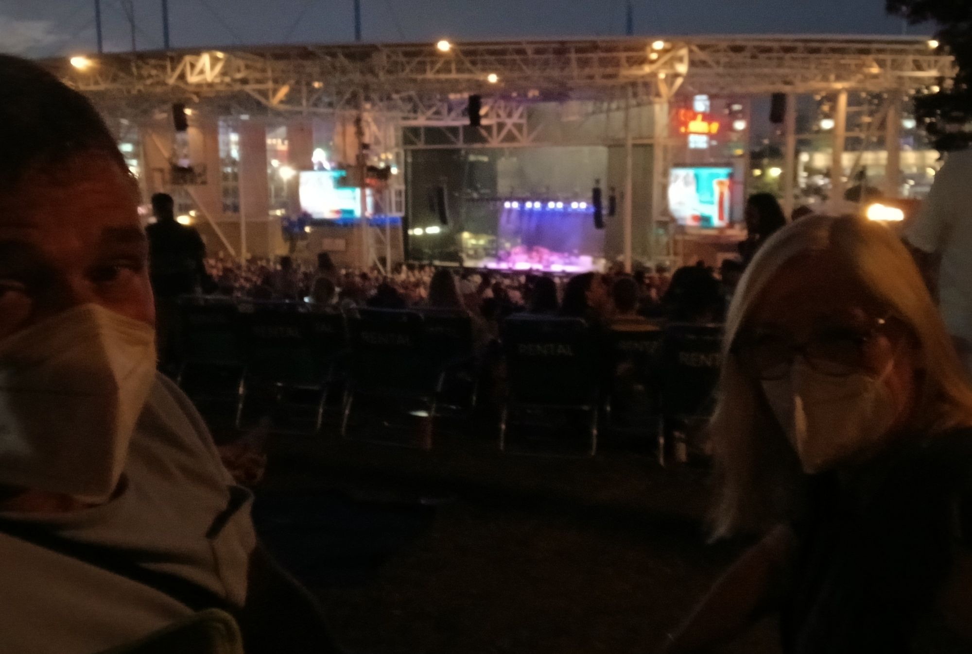 Kn95 masked couple sitting on lawn chairs at back of outdoor concert venue as night falls