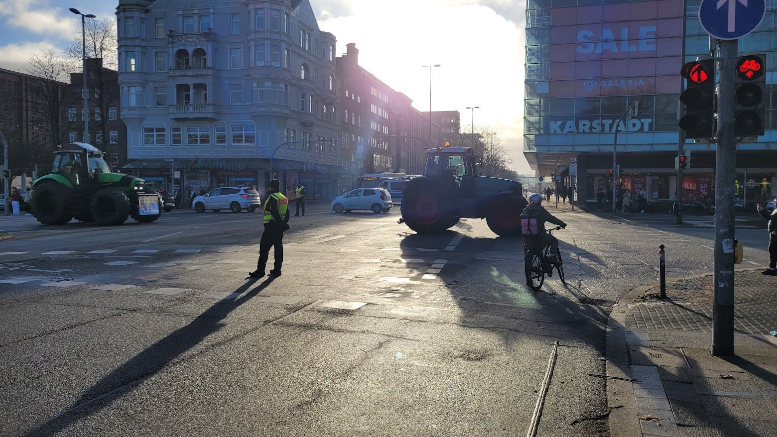 Straßenkreuzung, die von Polizisten abgesperrt ist. Von links queren Traktoren mit Plakaten an der Schnauze. Hinter der Kreuzung Links ein Mehrfamilienhaus, rechts ein Karstadt - Glaskasten. Die Sonne scheint von Oben durch die Straße, Menschen und Fahrzeuge werfen lange Schatten.