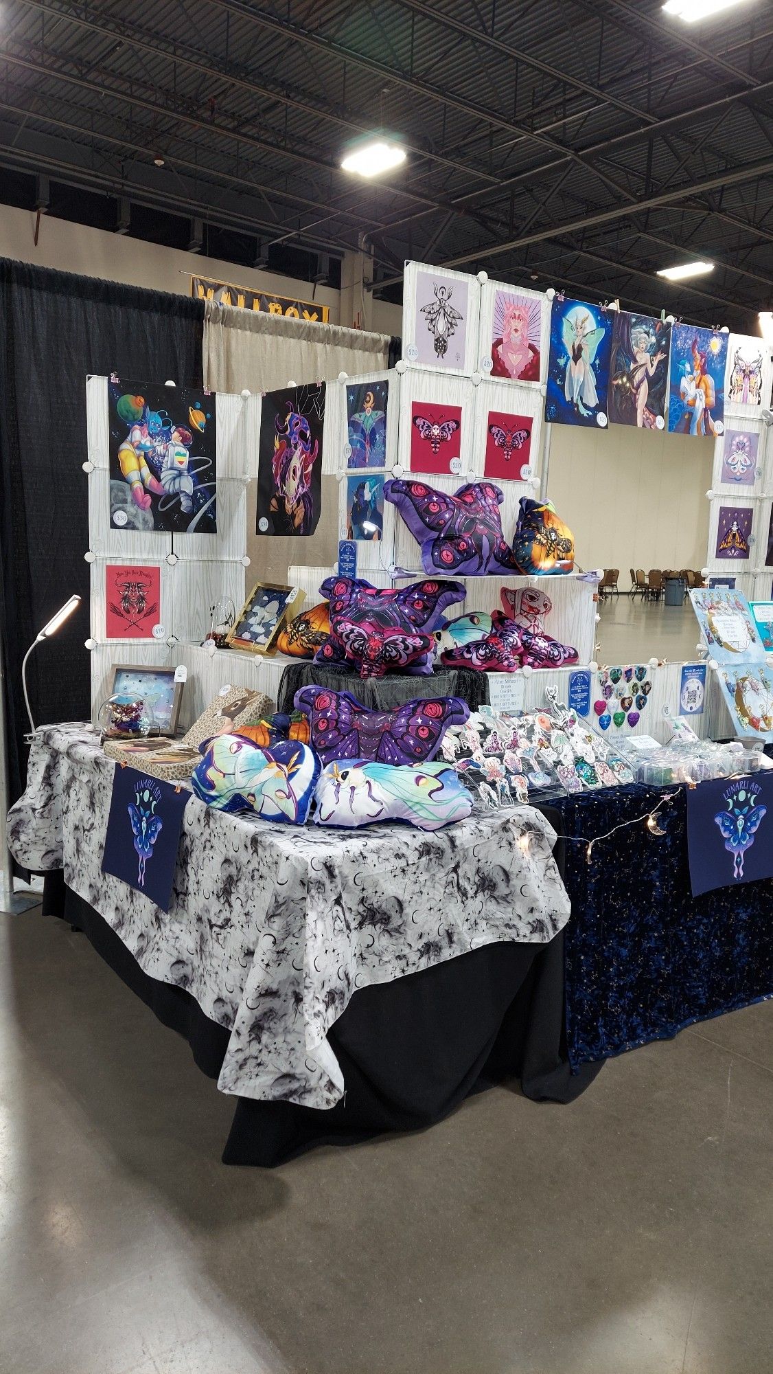 Three quarter view of the same setup which shows off the shelves of pillow plush. There are mothmen, luna moths, pumpkins with death's head and ghostly silk moths, pink death's head moth, and vampire pinup pillow plush. There is a black and white gauzy spiderweb fabric draping over the corner where rhe shelves join each other at a 90 degree angle at the corner of the table.