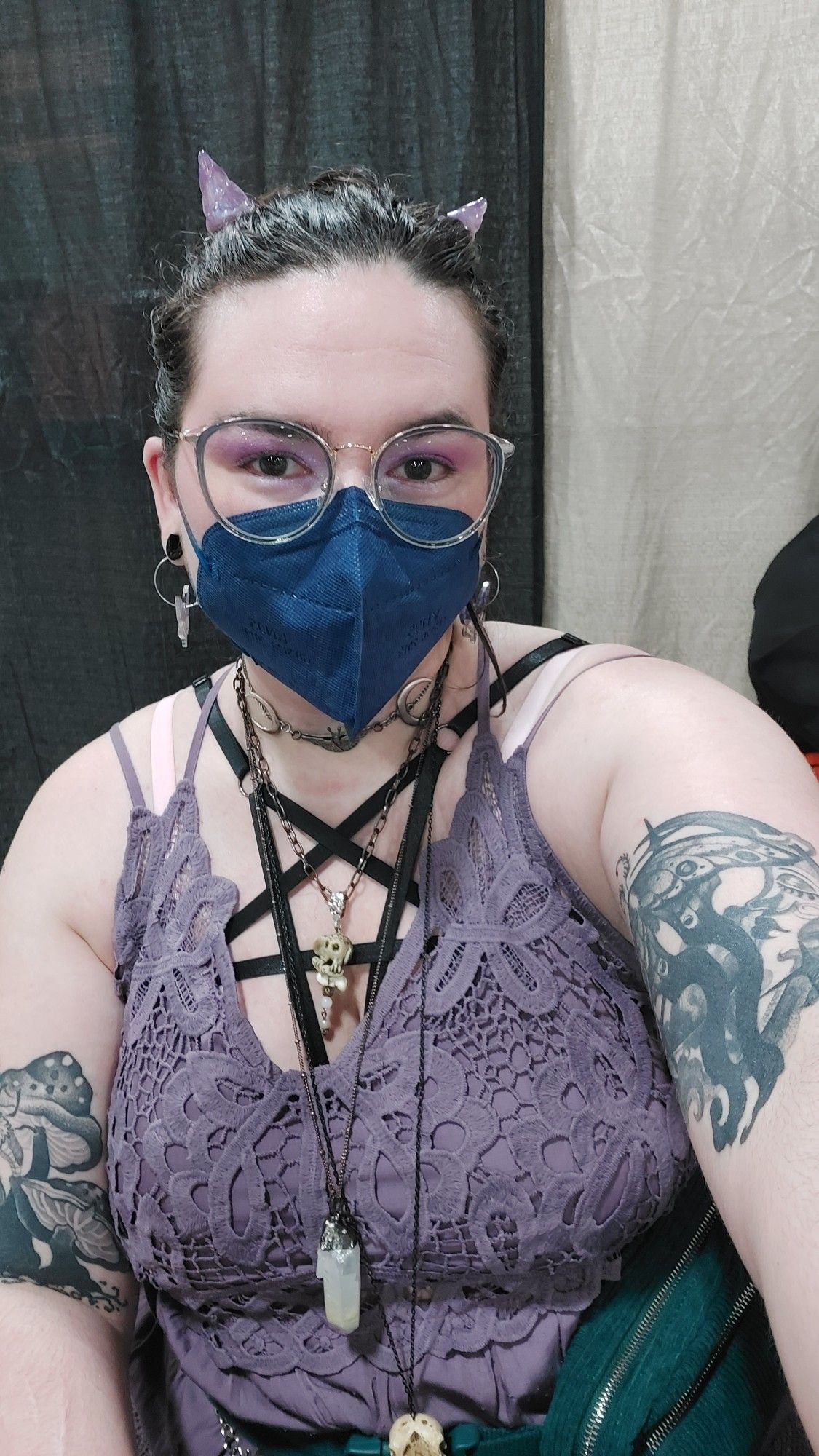 A selfish of me behind my table. I have brown hair pulled back, with 2 small demon horn clips that are shimmery purple and made of resin. I have purple eyeshadow and moth hoop earrings worn with black tunnels. I'm wearing a navy kn95 mask and have blue and gold glasses either people eyeshadow on. I am wearing a dusty purple laxy dress with a black pentagram harness underneath over my chest. I have 4 necklaces featuring moths, crystals, and faux bird skulls, and each of my arms has blackwork tattoos of moths, moonstone and mushrooms.