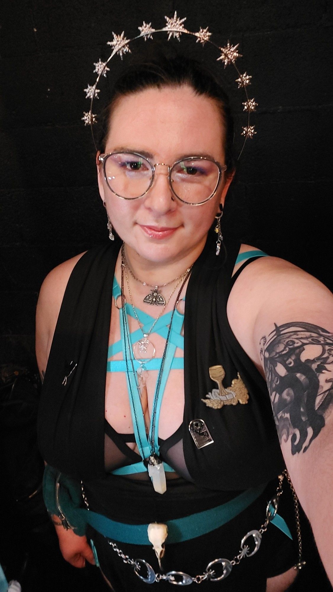 A selfie of wearing a plunge neck black wrap top with a mesh bra and bright blue harness underneath. I have on a long black, high slit skirt with blank hotpants and bright blue leg harnesses underneath. I am wearing 4 necklaces in various lengths, the longest of which is down to my belt. I have a faux bird skull necklace, a gunmetal necklace with an angel quartz crystal, a silver necklace with a star and butterfly, and a moth necklace. I am wearing a moon and star chain belt and a teal hip bag. I have a black and silver moth enamel pin and window enamel pin, and a silver and gold metal pin of a hand holding a goblet. I have dangling moon and star crystal earrings, blue and gold glasses, and a silver star halow crown. I have a blackwork tattoo of a crescent moon and luna moth visible on my arm that's holding out the camera. My hair is tied back and I have purple eyeshadow on. My eyes are very dark brown, my hair is brunette, and I have light skin.