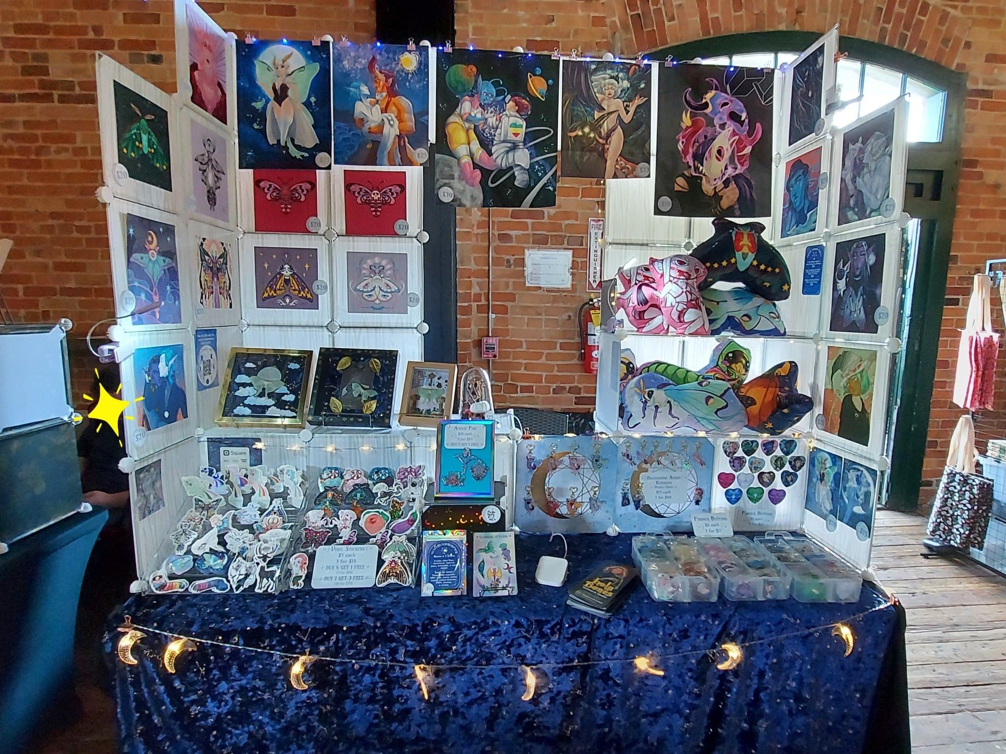 My 6' table display at a recent show. The venue has red brick interior walls and old wooden floors. My display has a black tablecloth, with a blue velvet table cloth with gold stars on top. There are crescent moon led lights strung across the front of the table. The display is largely made out of plastic grid cubes, with the back being 5 cubes high, and the side wings being 5 high on the inside and 4 high on the outside. Each panel is 12"x12". Many panels have prints fixed to them for display, with themes of moths, original fantasy characters, and one critical role print. There are matte and holographic prints. 

One side of the display has shelves built in to display pillow plush. The other aide has shelves to display shadowboxes with moth specimens. On the table in front of the display are clear racks for stickers, and displays for charms and buttons. There are also acrylic pins propped up in a holographic frame, and mystery sticker packs and sticker sheets on wire easels.