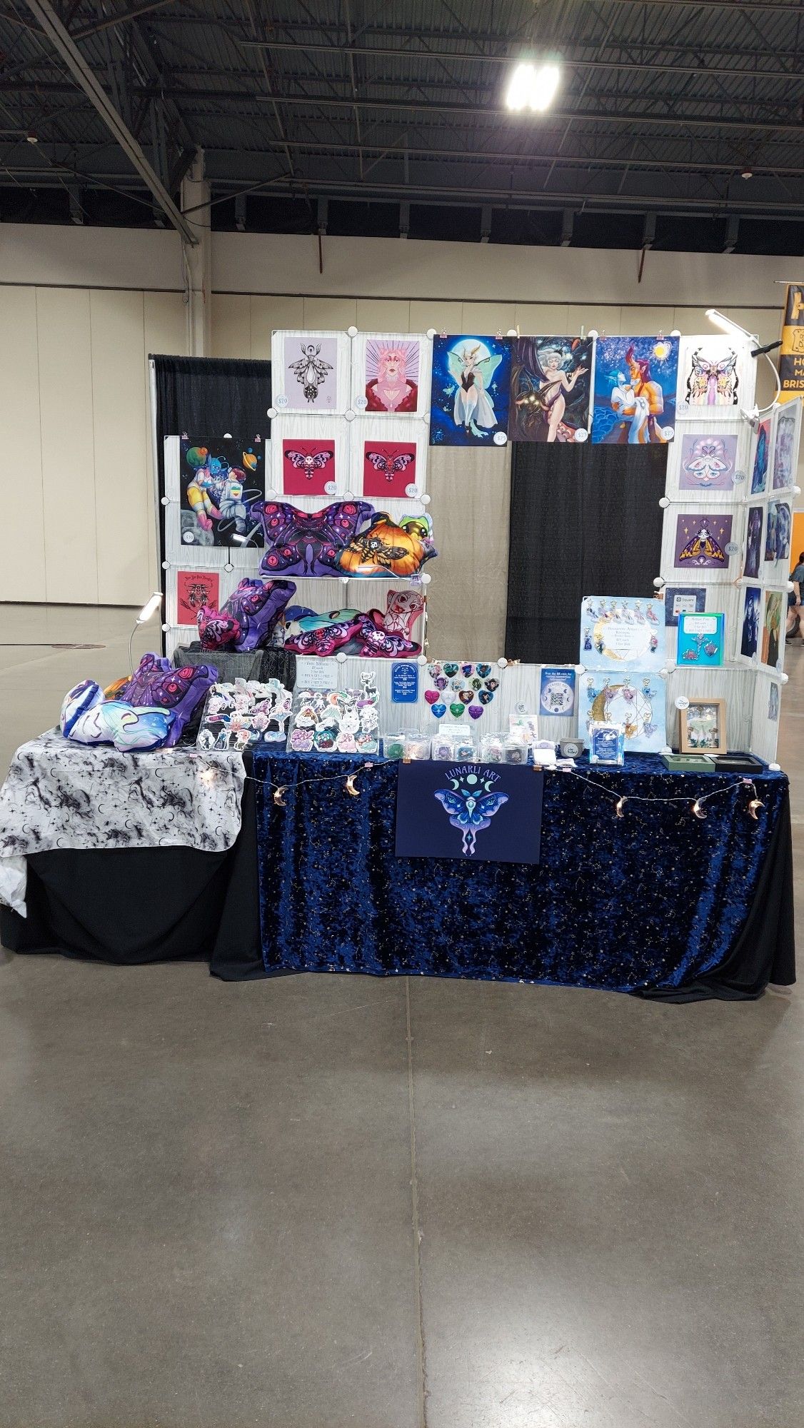 The front of my artist booth setup for World Oddities Expo. There are two six foot tables pushed together in an L shape, laid out so customers walk around the perimeter of the L. Both tables have a black cloth underneath with a decorative cloth on top. One table has a black and white cloth with mysterious smoke and moons as a pattern. Another is deep blue velvet with shimmery gold star and moon details. The structure on top or the tables is comprised of plastic sheet grid cubes snapped together to form walls with openings and intermittent boxes. There are prints and buttons displayed adhered to the cubes, and pillow plush on the sure shelves and tables. There are stickers in clear plastic racks. There is a frame for pins on another shelf and 2 panels with charms. There is a sign thay says Lunarli Art with my moth logo.