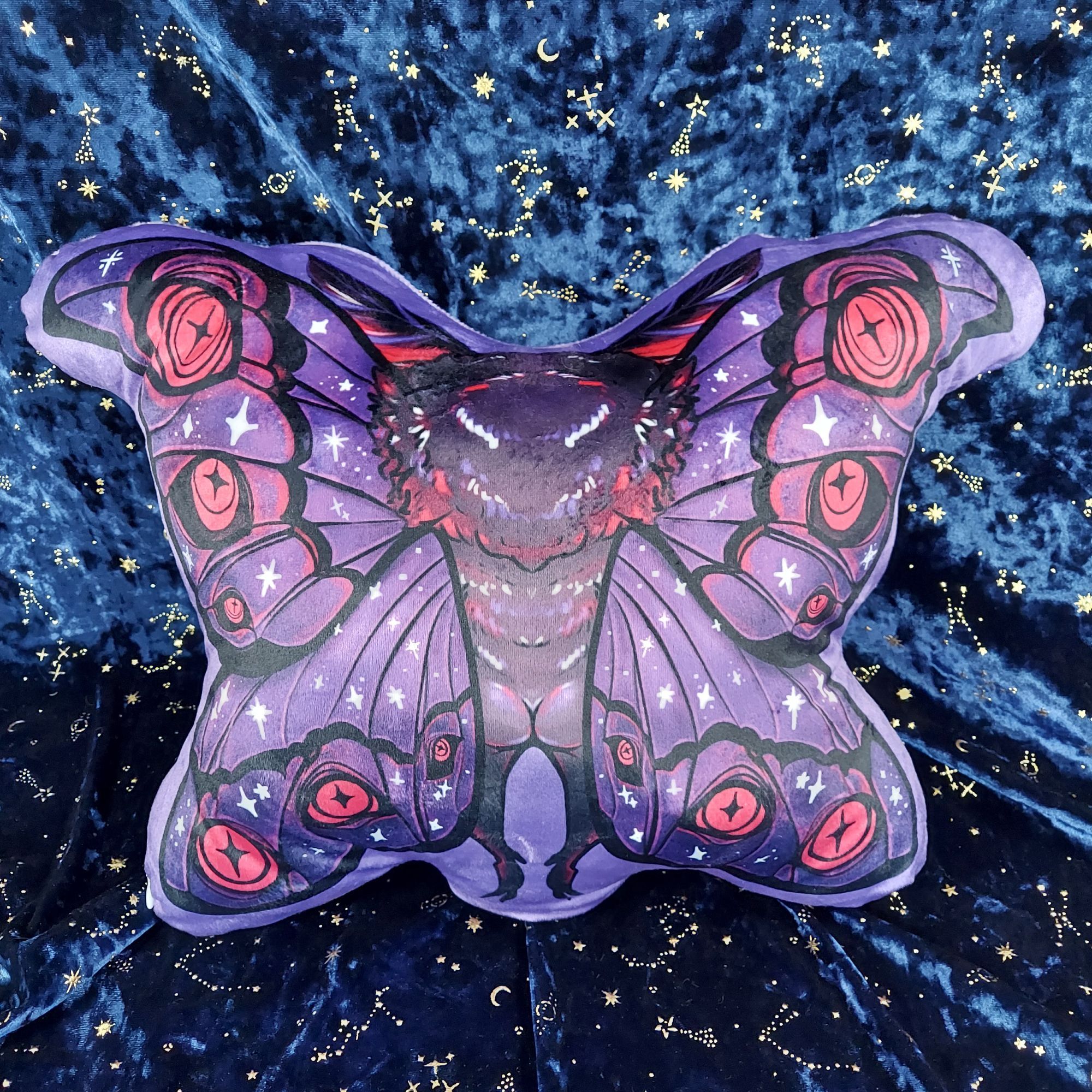 The back of my Mothman fairy pillow plush, displayed against a dark blue velvet background embellished with shimmery gold stars and moons. It is printed on a velveteen fabric that has a slight shine from the lighting. He is puffed up with ample stuffing and he looks CHONKY. From the back of the design, you can see he has a very round, perky booty. The back view of his head is drawn to be very fluffy. His arms are not visible from the back since they are obscured by his large wings, which have the same design on the back as the front.