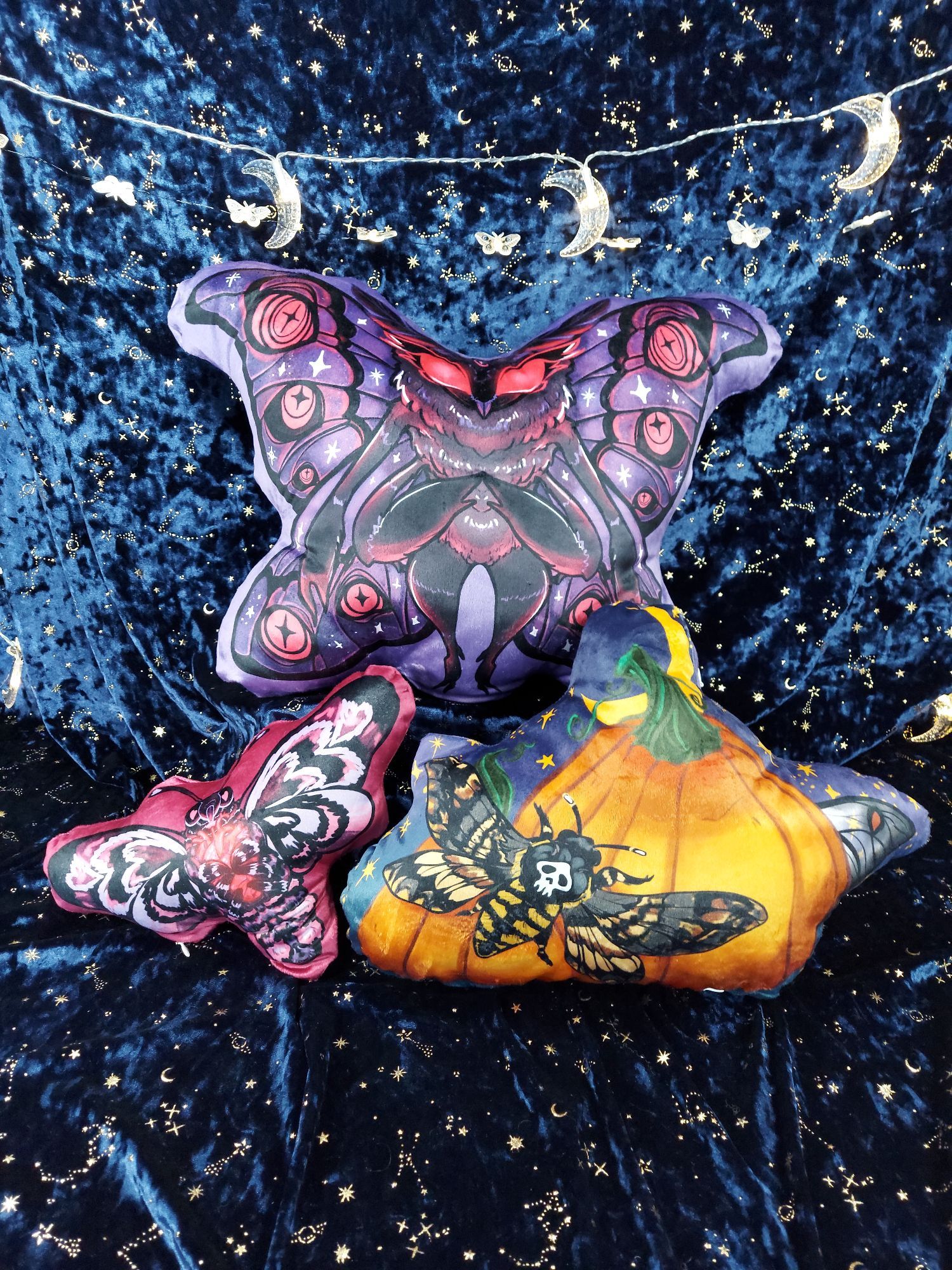 3 shaped pillow plush displayed against a blue velvet backdrop with shimmery gold moons and stars. There are moon and moth-shaped LED string lights above them. On a riser above and behind the other 2 pillows is a giant Mothman fairy pillow. He is black, purple, red, and dark grey. He has big red eyes and a barn-owl like face with moth-like wings, body, and arms. The two pillows in front and just below him are a pink death's head moth pillow and a pumpkin & moths pillow. The pink & black death's head moth is holding a glowing anatomical heart. The other pillow is 2 orange pumpkins, with a naturally colored death's head moth resting on 1 pumpkin. You can see the wing-tip of a ghostly silk moth peeking out from the opposite side There is a bit of a starry night sky with a yellow crescent moon behind the scene. All pillows are a velveteen fabric, and shaped in a silhouette to the art, so they are all organic, non-rectangular shapes.