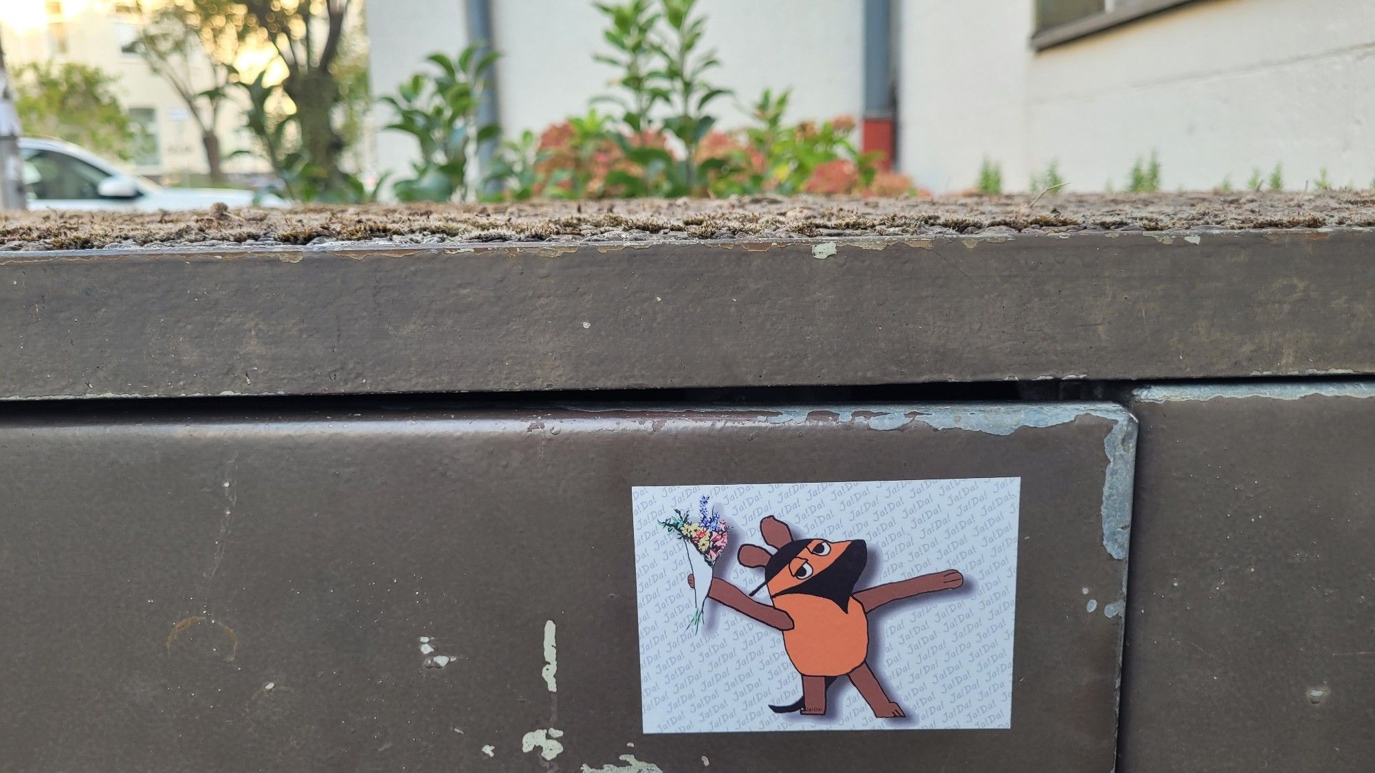 Obere Kante von einer Mülltonnenverkleidung. Darauf ein Aufkleber auf dem die vermummte Maus (orange) einen Blumenstraß wirft (vgl. Banksy). Im Hintergrund etwas grün.