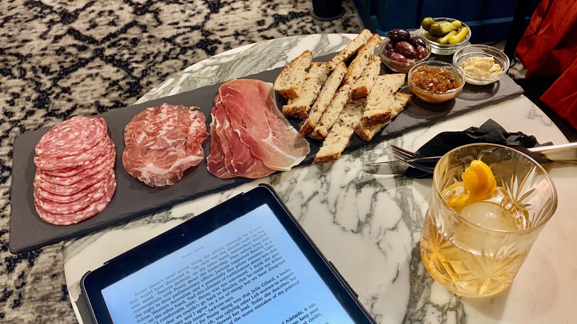 On a small round marble table sits a large charcuterie board containing a generous serve of three kinds of cured meat, pieces whole grain bread, and small bowls of Kalamata olives, gherkins, chutney, and perhaps the last one is mustard? In a lowball glass is an old fashioned. And on an iPad is Margaret Simons’ biography of Penny Wong.