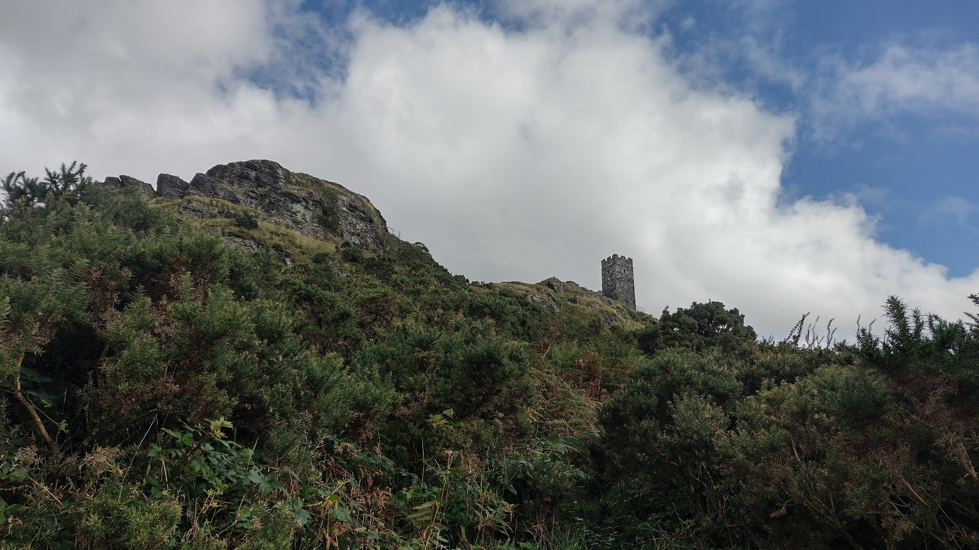 Upward view of a rocky outcrop with a church tower at the top, against a cloudy blue sky.