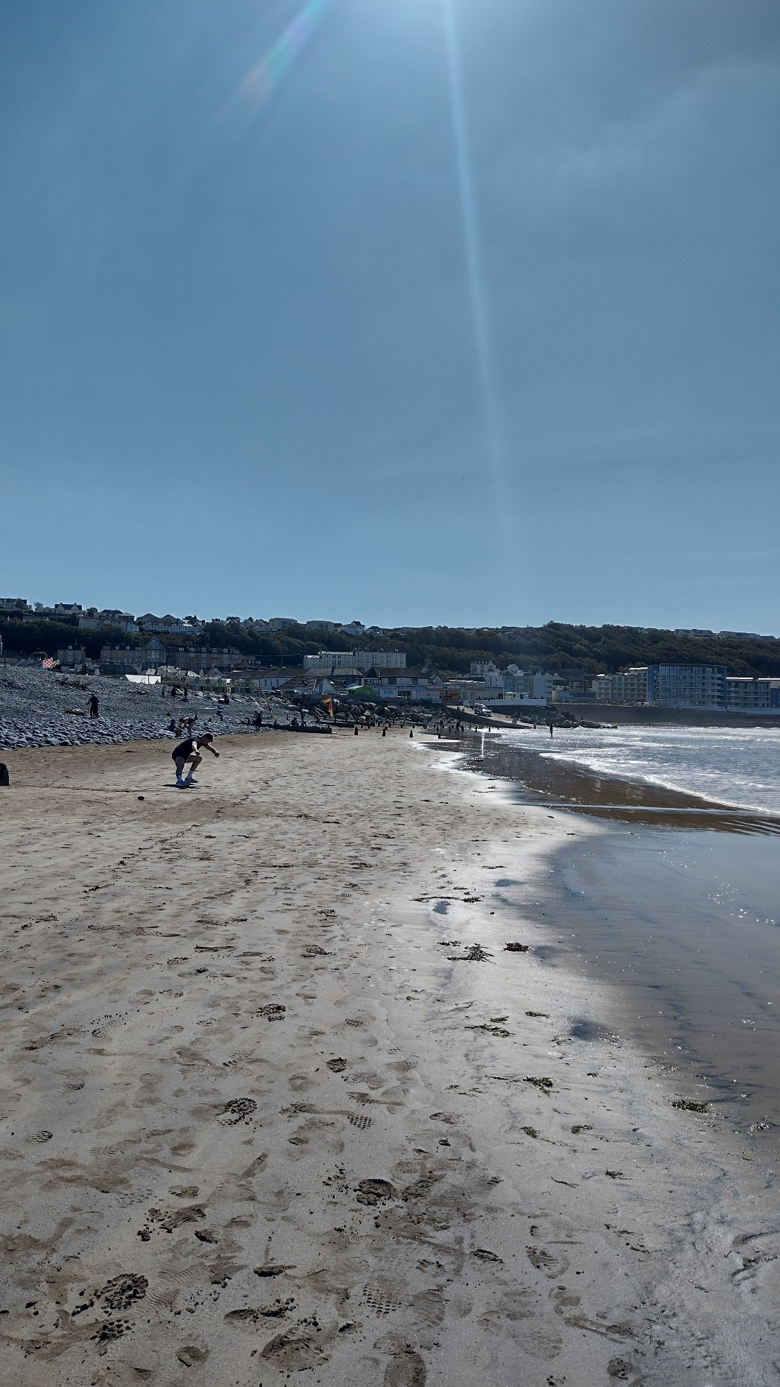 Image along a beach with sunlight shining on the water.  There are hills and buildings in the distance against a clear sky.