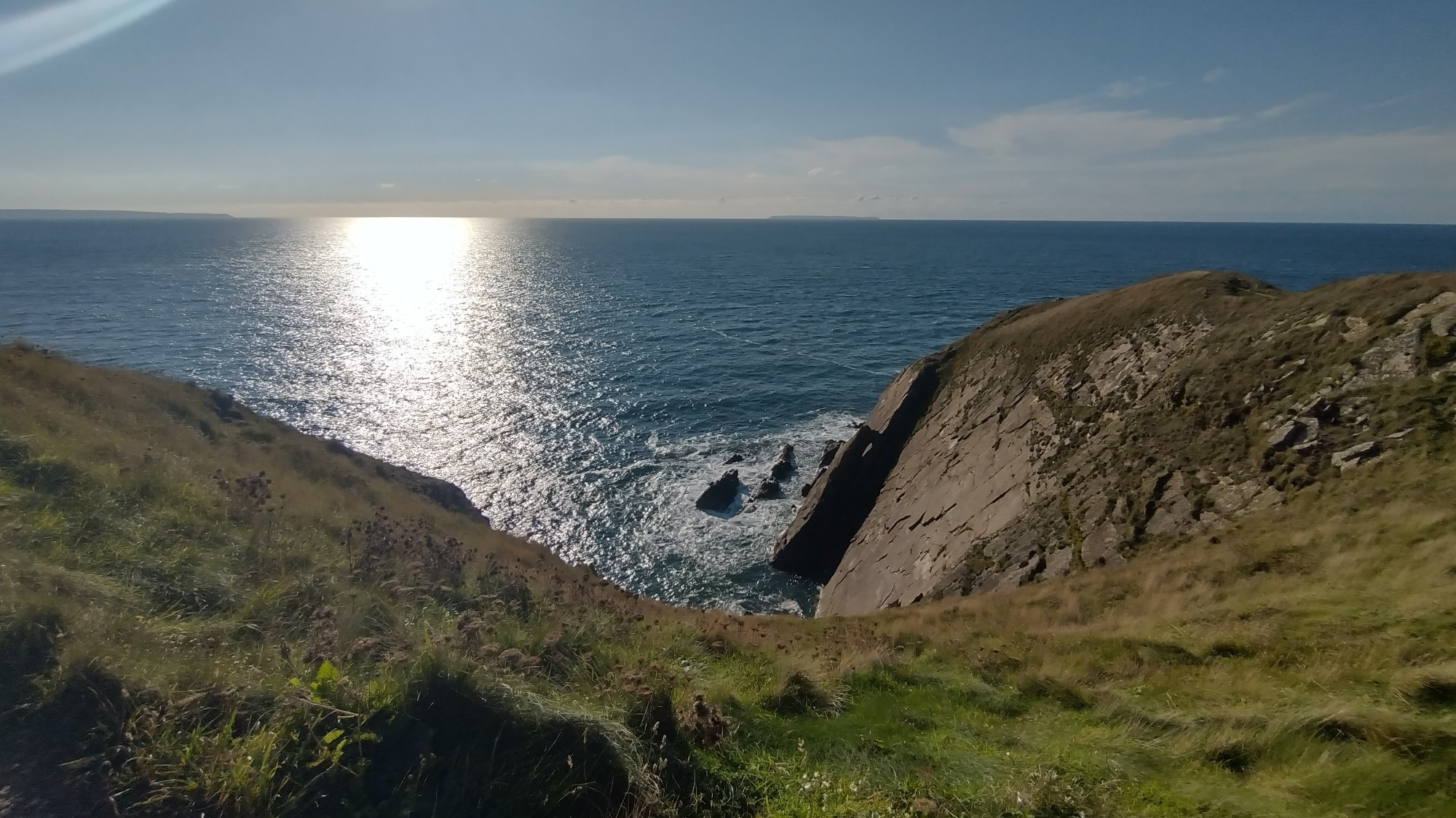 Image of a cliff face with rocks tilted upwards in the sea.  The sun is shining on the sea surface.