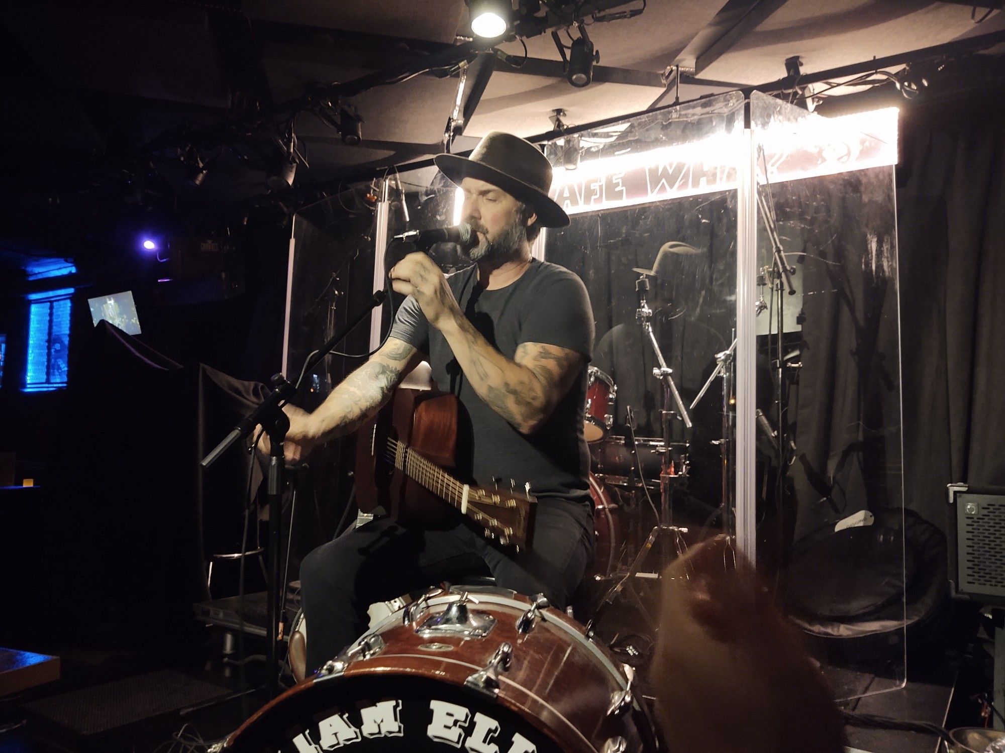 Folksinger William Elliott Whitmore on stage about 6 feet from us. He's adjusting the microphone with an acoustic guitar balanced on his thigh. A bass drum is in front of him and part of his name is visible on it. He's dressed in black pants, black T-shirt, and a brown hat. He's got a salt and pepper beard and his arms and hands are heavily tattooed. My husband's clapping hands are visible in the lower right corner of the picture.