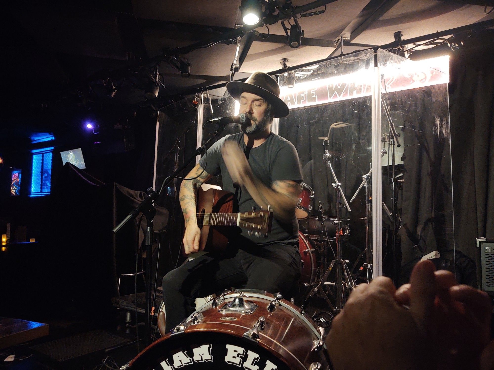 Folksinger William Elliott Whitmore on stage about 6 feet from us. He's adjusting the microphone with an acoustic guitar balanced on his thigh. A bass drum is in front of him and part of his name is visible on it. He's dressed in black pants, black T-shirt, and a brown hat. He's got a salt and pepper beard and his arms and hands are heavily tattooed. My husband's clapping hands are visible in the lower right corner of the picture.