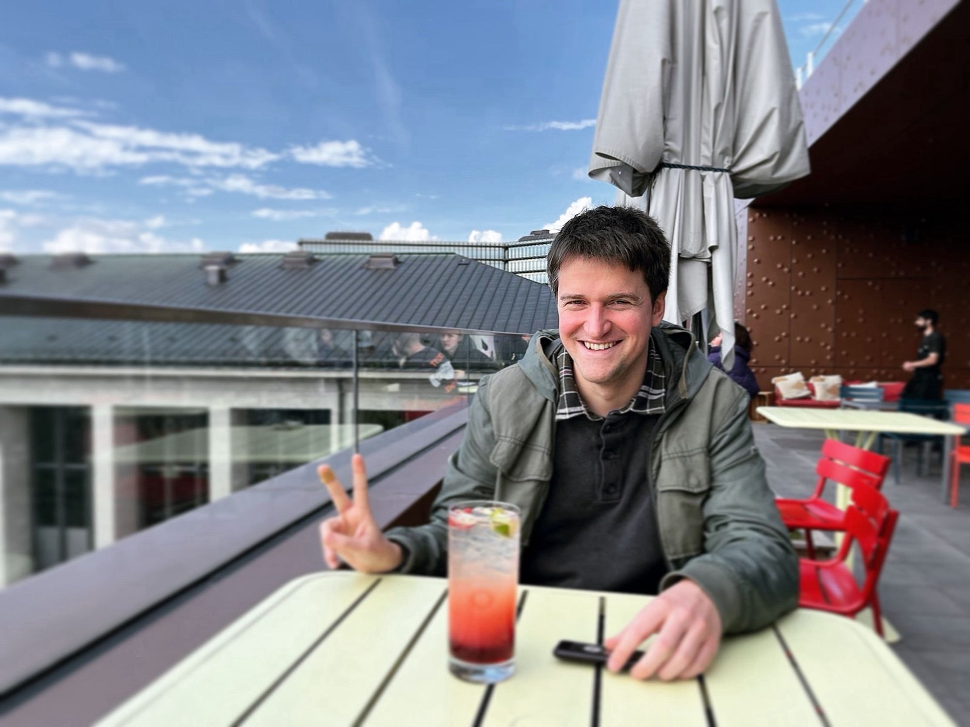 A picture of me sitting at the rooftop café “Frau im Mond” atop the German Museum in Munich. Clear blue sky and clouds. The table is one of those crappy metal cafe tables and is light green. The chairs are all red. 
I’m smiling and showing the peace sign with my right hand. There is a bandaid on my finger. In front of me is the signature nonalcoholic house cocktail, deep red at the bottom and transparent at the top, which set me back 6 euros. It was good though. In my left hand, the food buzzer. In the background, the waiter looks off longingly into the distance. Don’t worry buddy, it’s the weekend soon. TGIF.