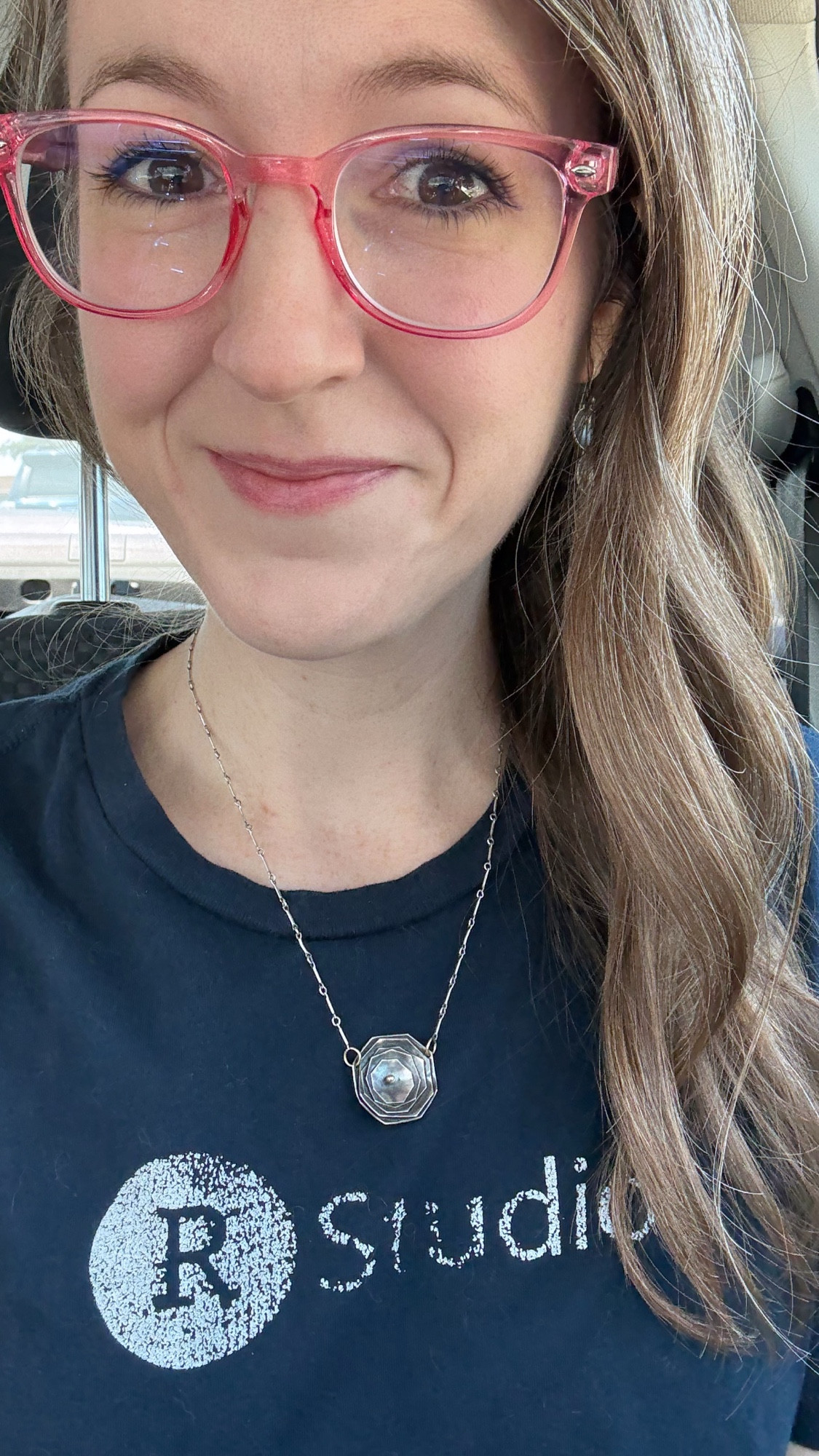 Libby, sitting in her car, wearing a navy blue t-shirt that says RStudio on it. The printed logo is flaking and rubbed off. Libby is a smiling woman with light brown hair, brown eyes, pink glasses, and a silver necklace.
