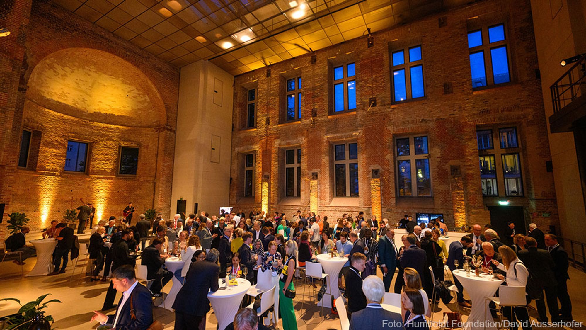 A hall can be seen from above, in warm light, with many people standing at tables and chatting.