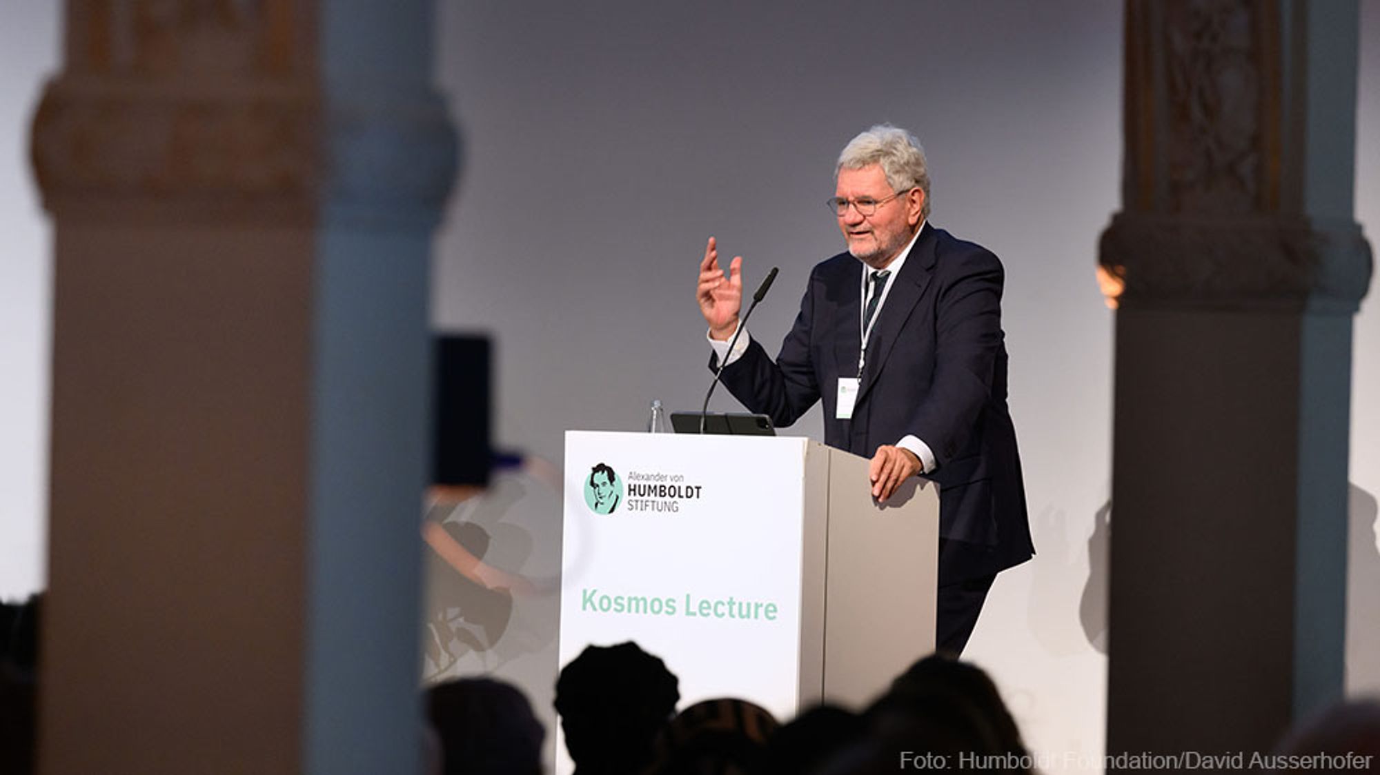 A man stands behind a lectern and speaks to the crowd. It says "Kosmos Lecture" on the lectern.