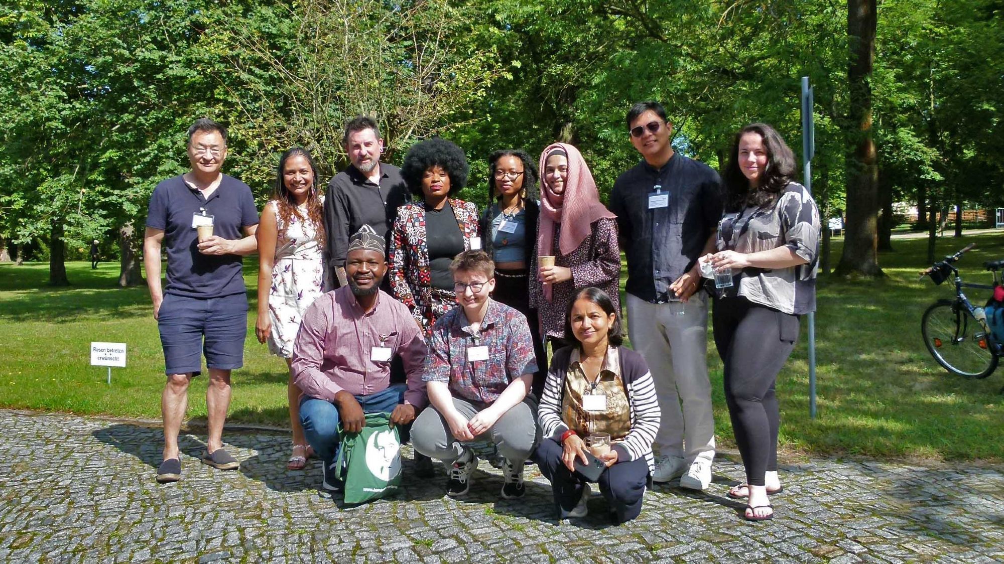 Group picture of eleven people smiling at the camera, behind them  a lawn with trees