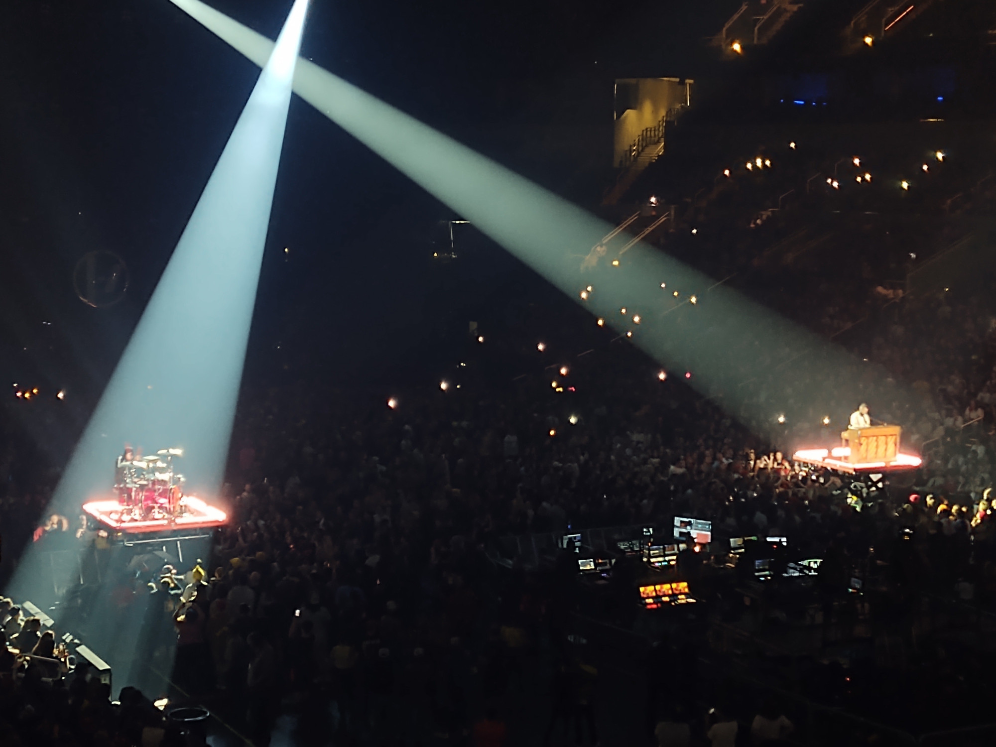 A stadium filled with concert goers for Twenty One Pilots. Josh Dun sits on the left, in the pit, with him and his drumset illuminated by a spotlight. Tyler Joseph sits on the right, in the pit, with him and his piano illuminated in spotlight.