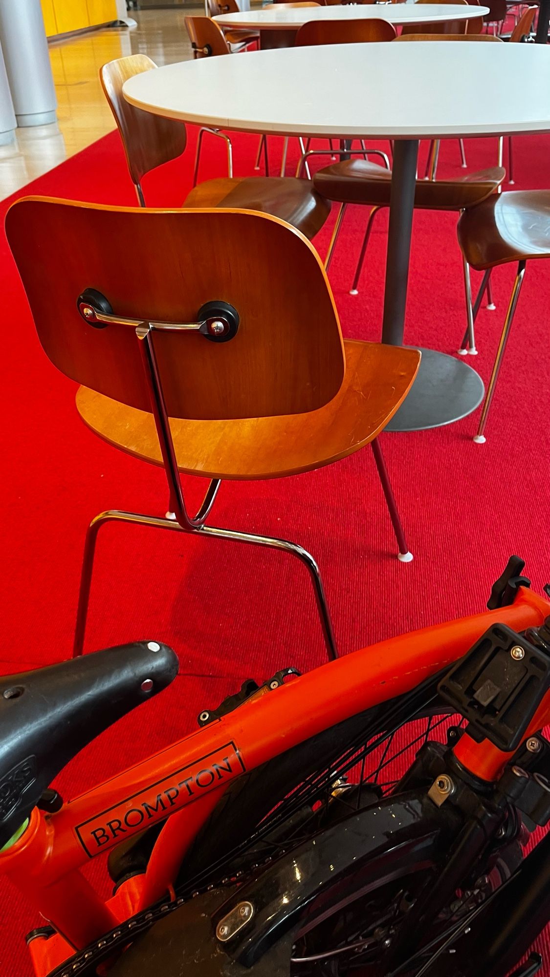 An orange Brompton bicycle is parked beside a round white table with wooden Eames chairs in a space with red carpeting.