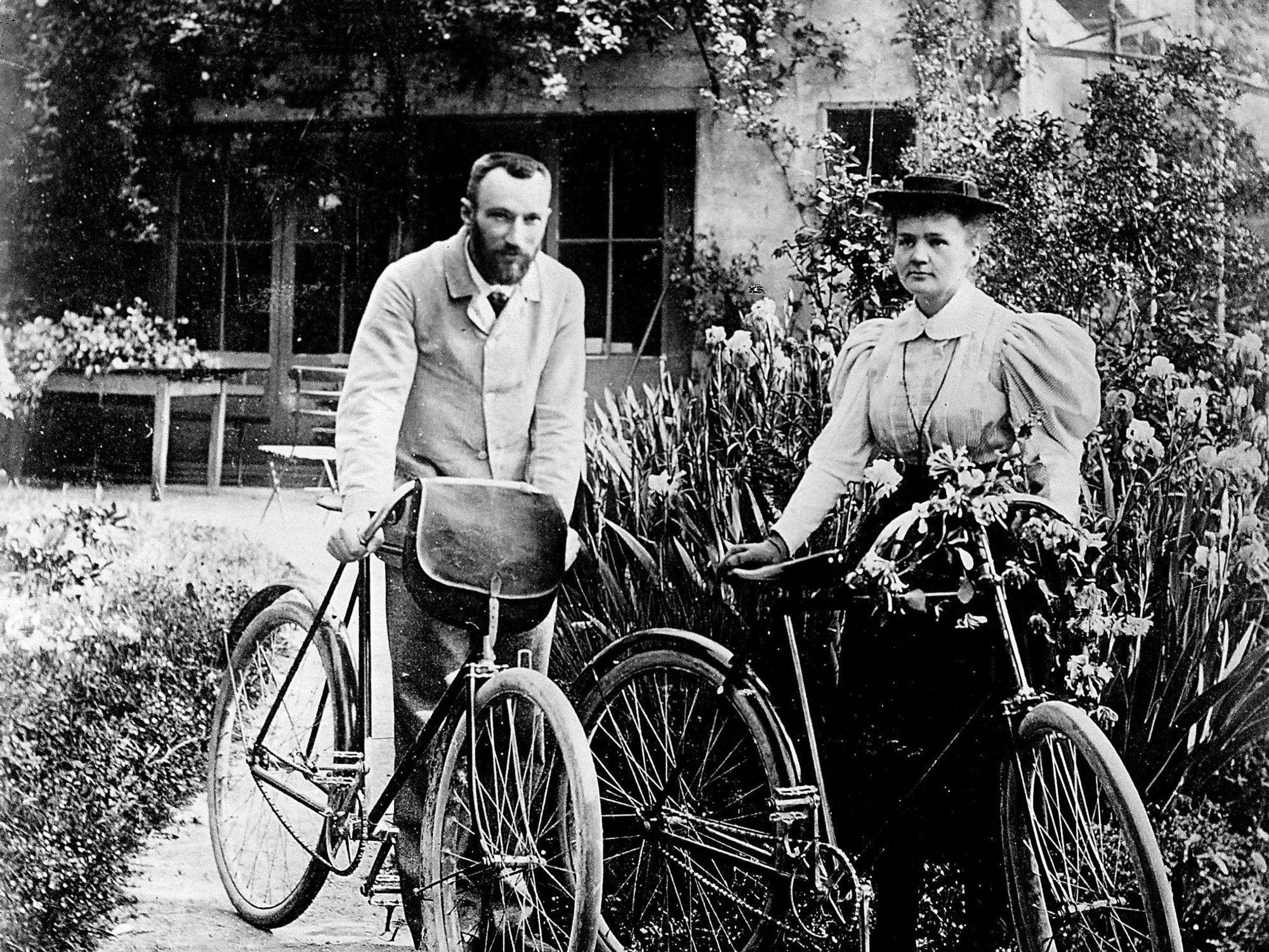 A black and white photograph of a man and woman standing beside their bicycles in a garden. The man is wearing a light suit and has a beard, while the woman is dressed in a stylish, puffed-sleeve blouse and holding a bicycle adorned with flowers.