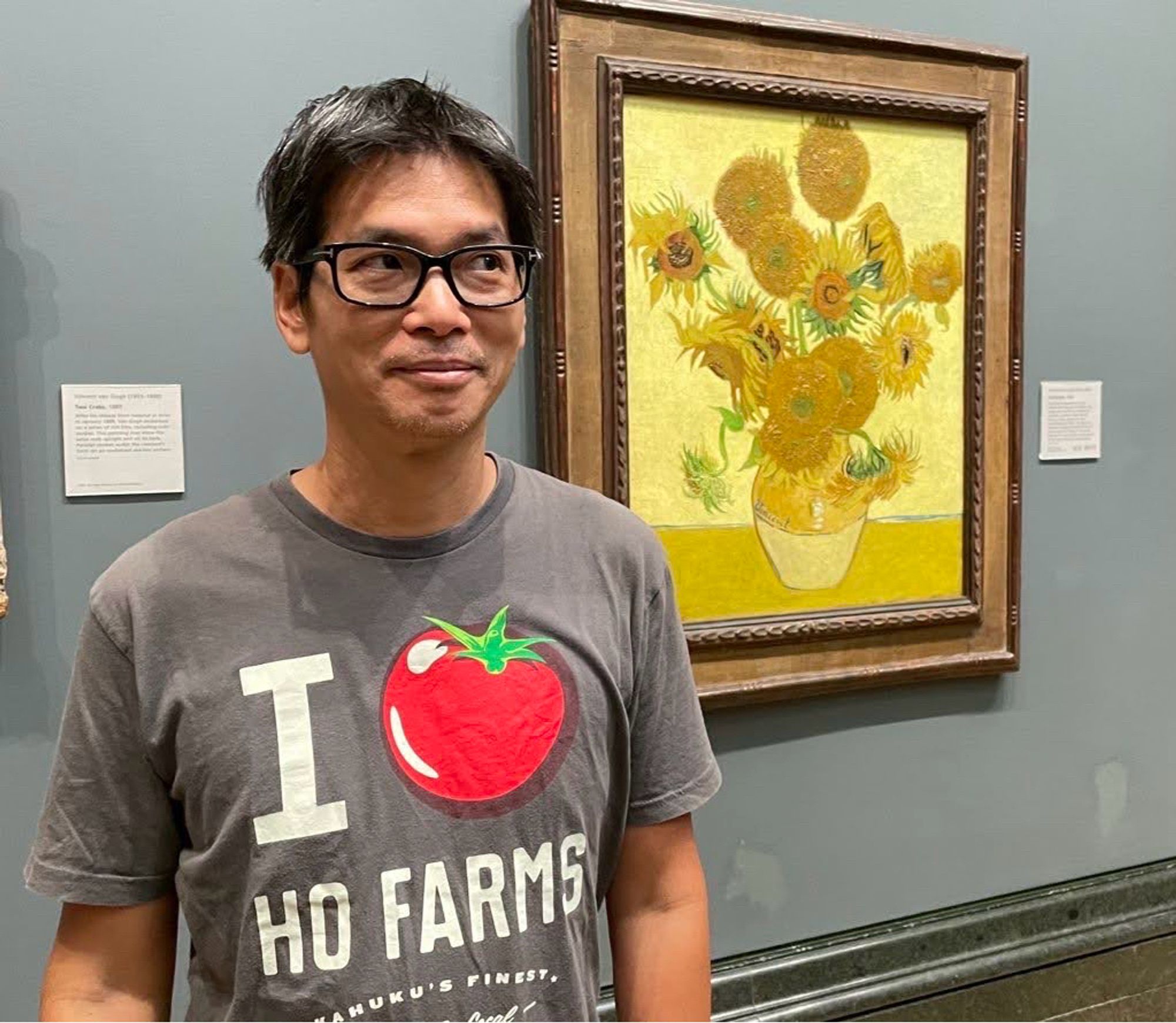 A person wearing a gray shirt with a tomato graphic stands in front of Vincent van Gogh's famous "Sunflowers" painting. The wall behind them is painted gray, and there are museum labels nearby.