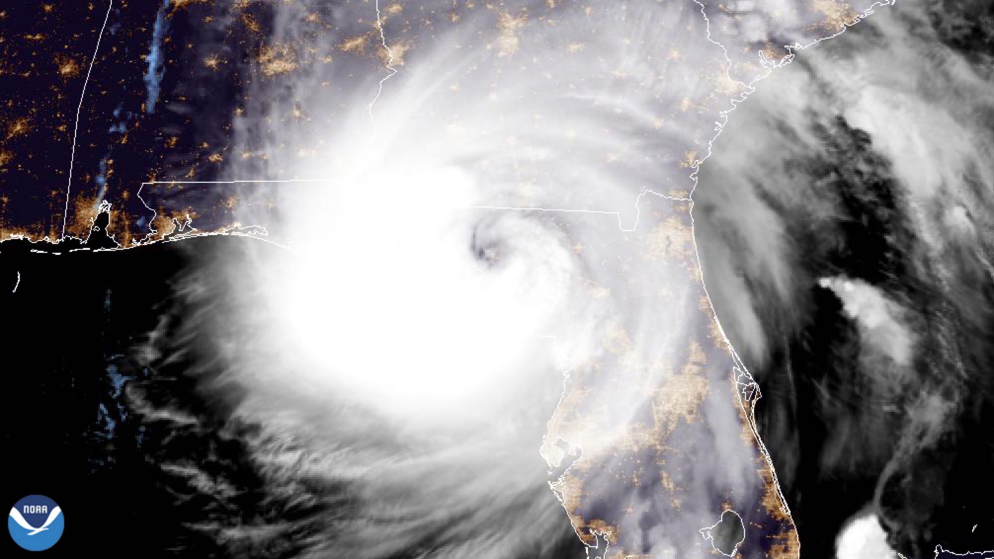 Satellite imagery of a hurricane approaching the southeastern United States, with a spiral cloud structure and bright, intense center. Surrounding areas are illuminated, indicating land features and urban regions.